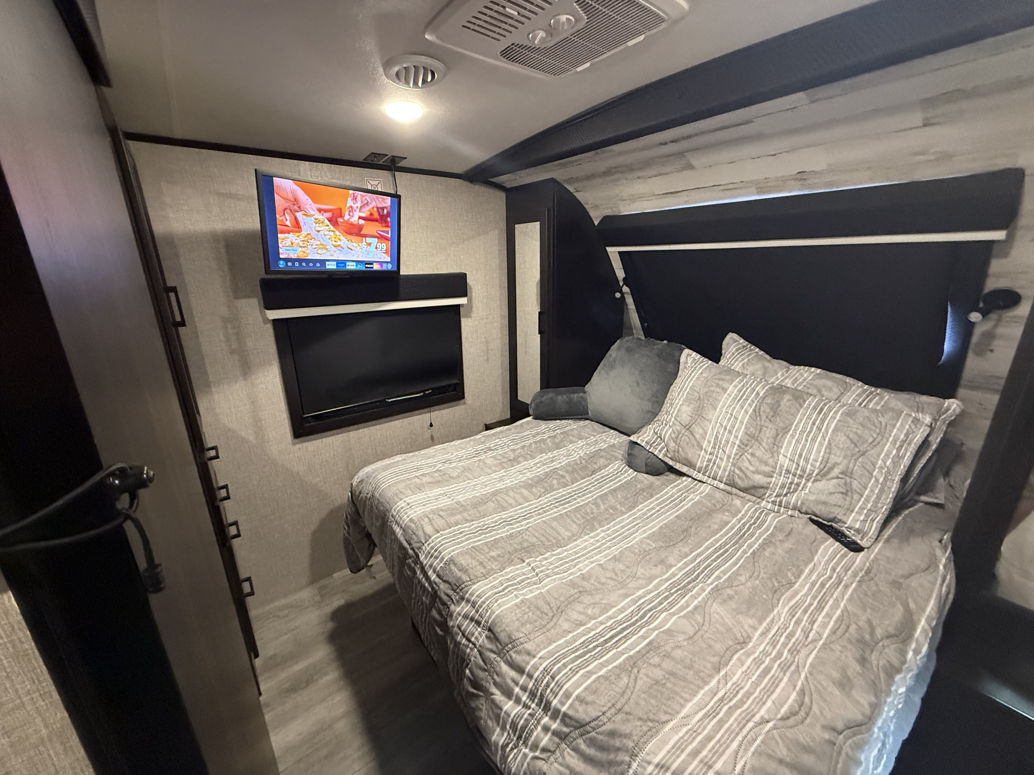 Cozy RV rental bedroom featuring a striped bedspread, mounted TV showing a cooking show, and overhead lighting. Ideal for travel.. Jayco White Hawk 2023