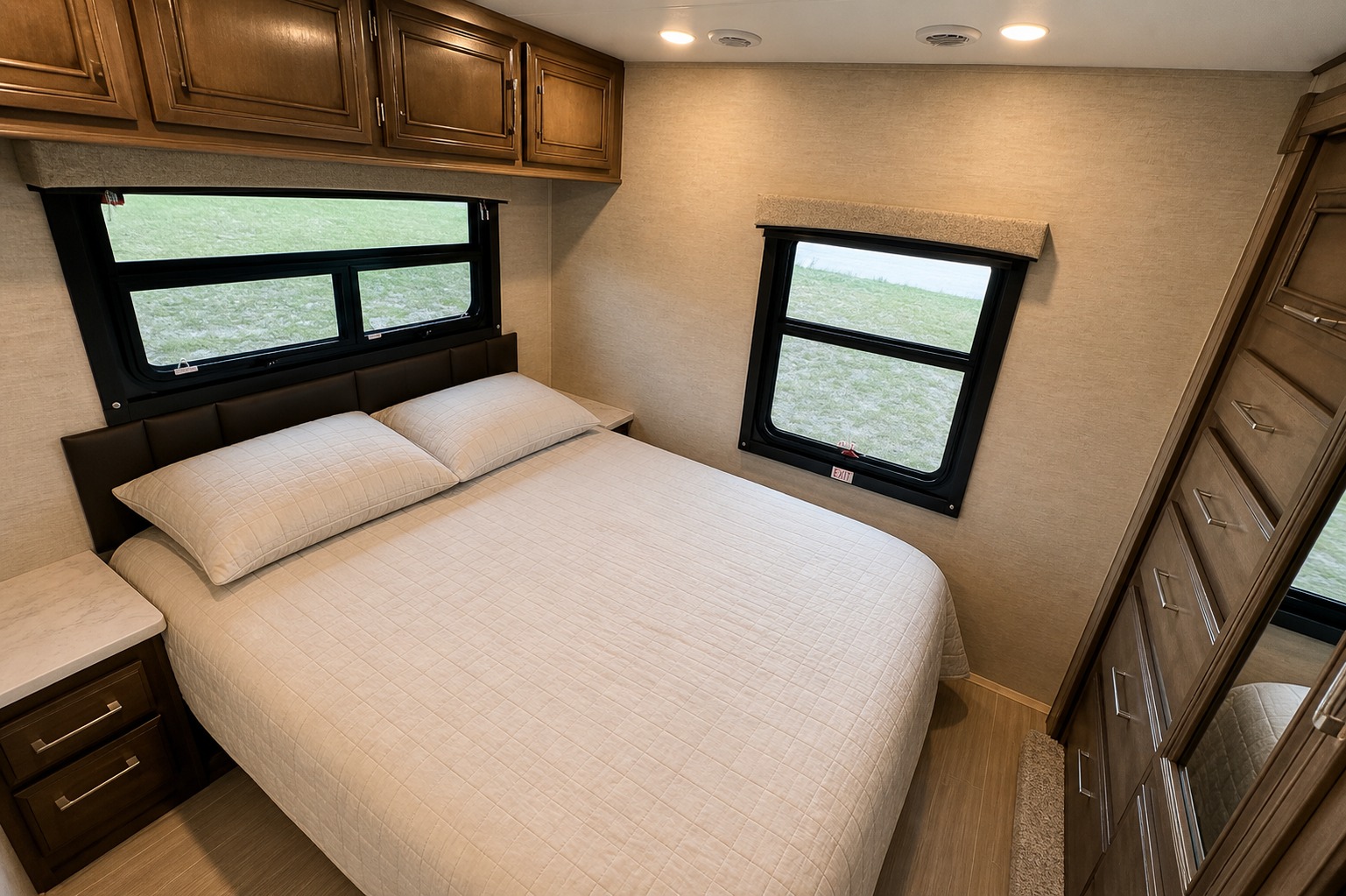 Cozy RV rental bedroom featuring a comfortable bed, large windows looking out to a green field, overhead cabinets, and a tall dresser.. Entegra Coach Odyssey 2023