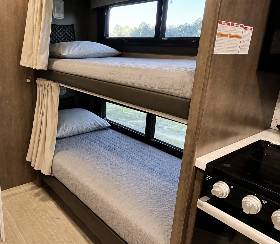 RV rental interior: stacked bunk beds with light gray bedding & window views. Stove on right. Ready for your family trip!. Entegra Coach Odyssey 2023