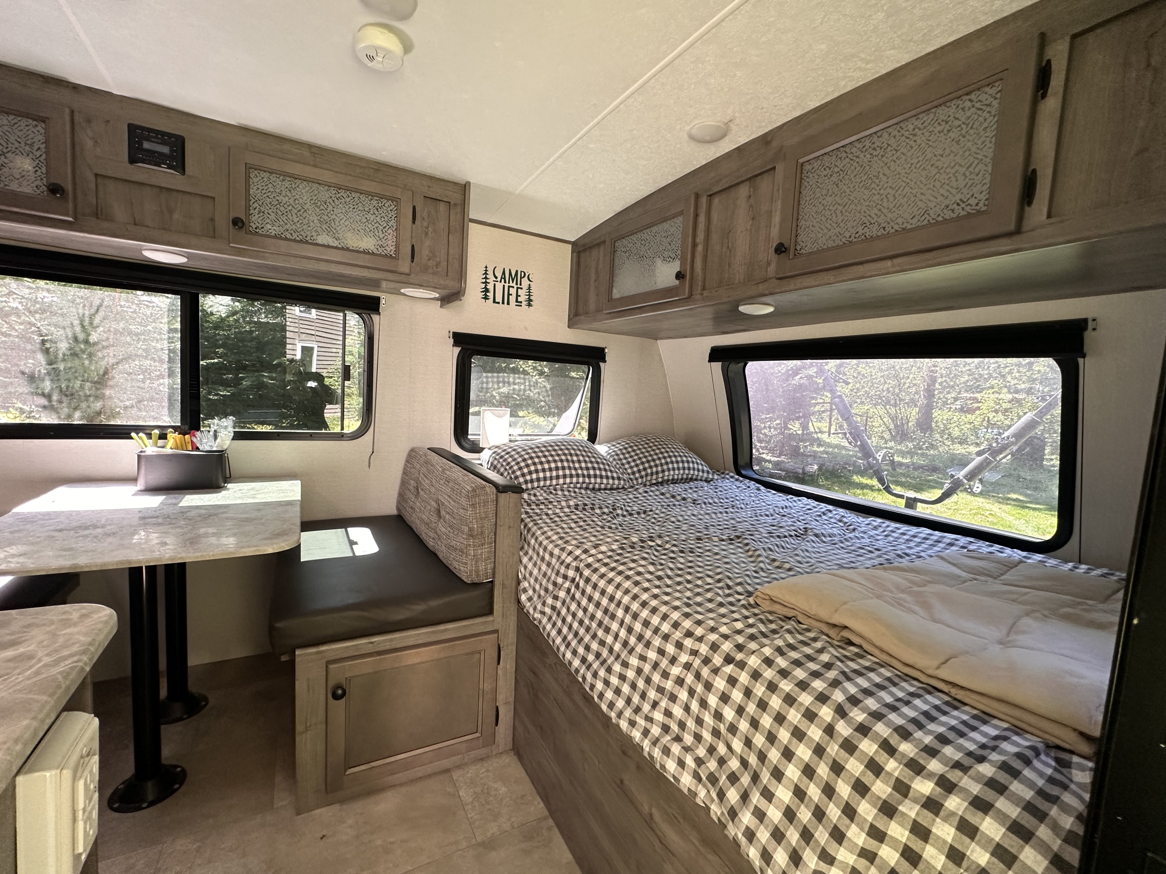 Cozy rv rental interior with a gingham bed, dinette, overhead cabinets, and large windows showcasing the outdoors.. Coachmen Apex Nano 2021