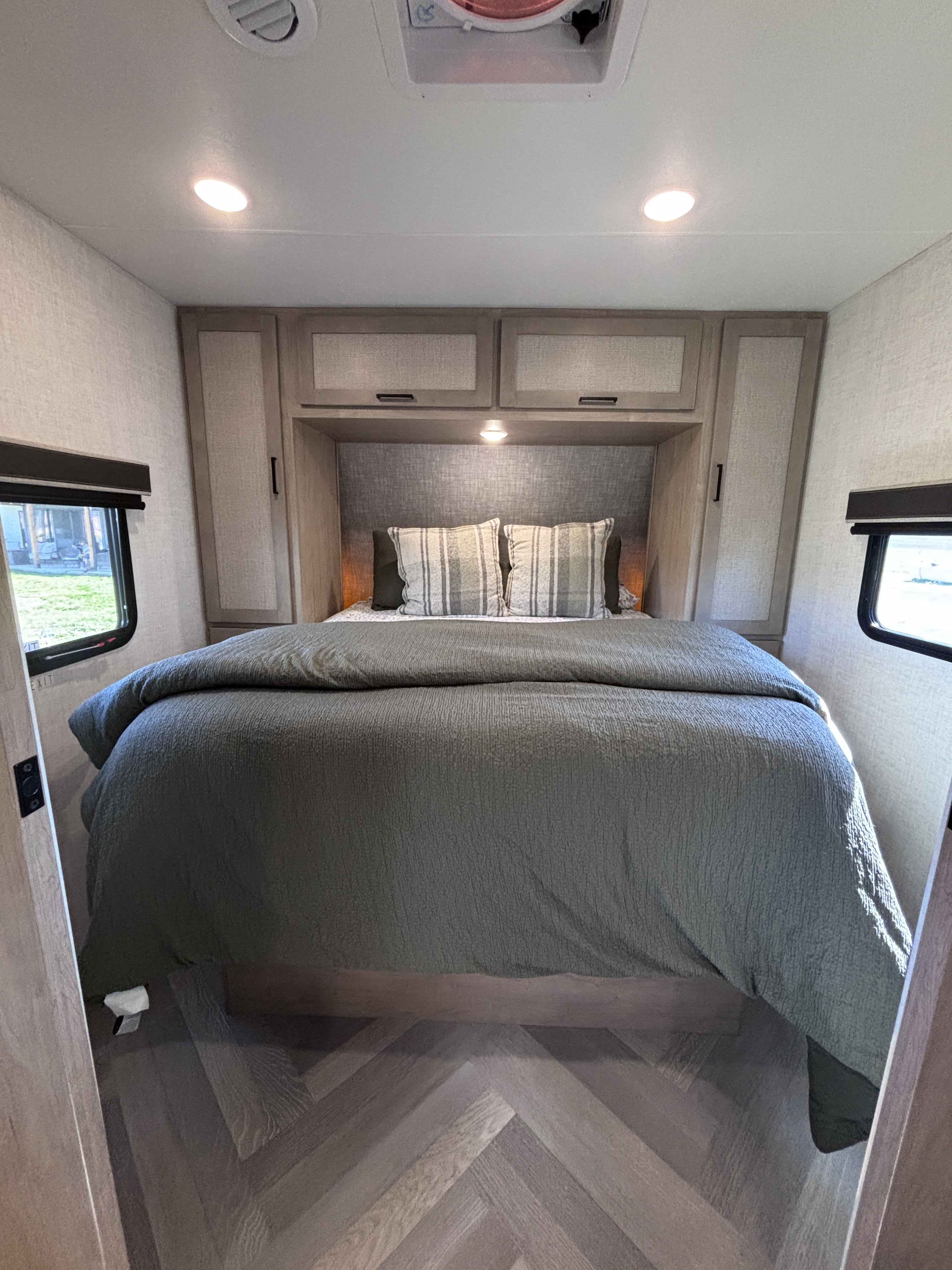 Inside an inviting RV rental bedroom featuring a bed with a textured green comforter, overhead storage, and herringbone flooring.. Ember E 2025