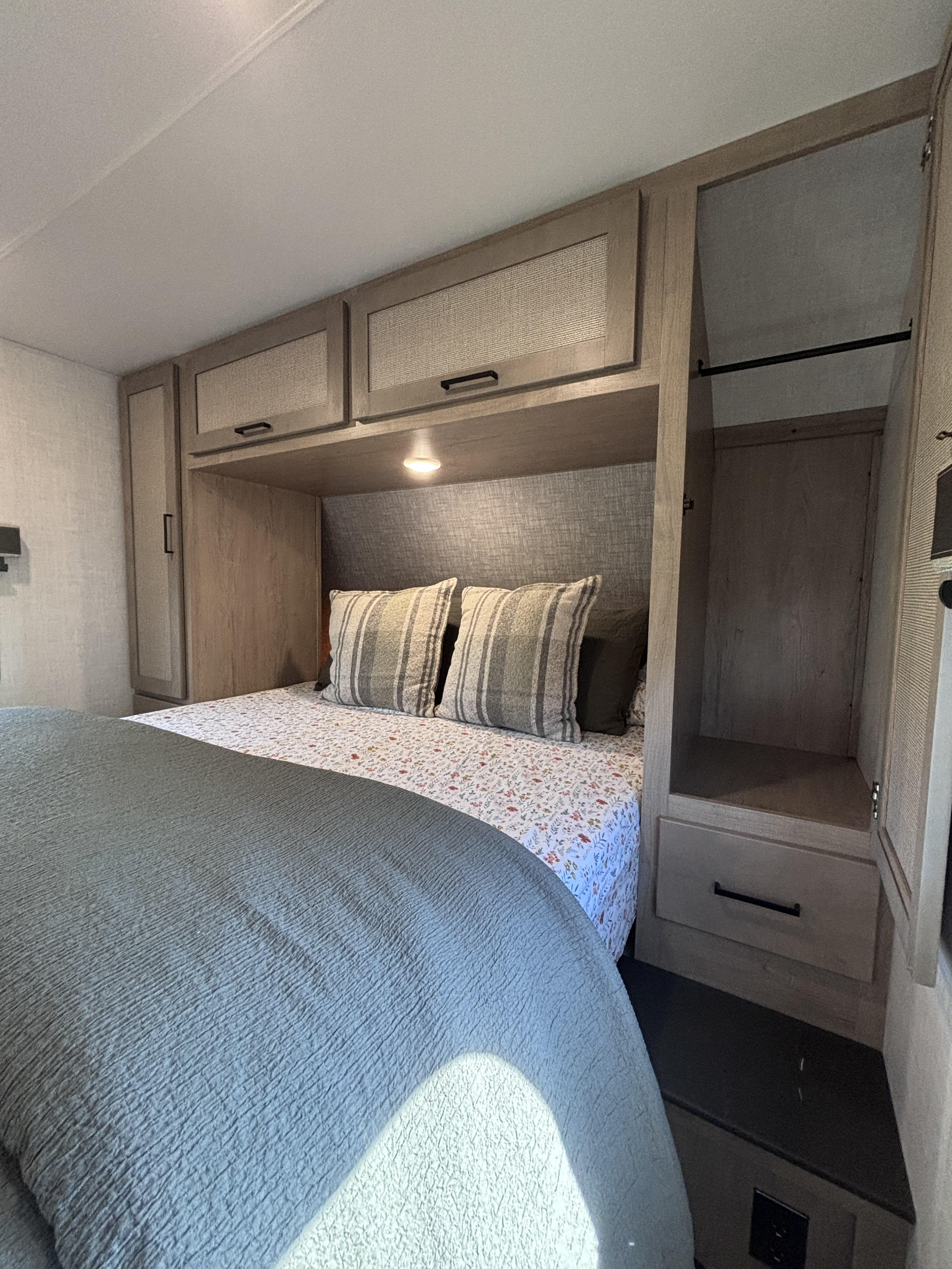 Cozy RV rental bedroom featuring built-in storage cabinets above a bed with floral sheets and striped pillows, plus an open wardrobe.. Ember E 2025