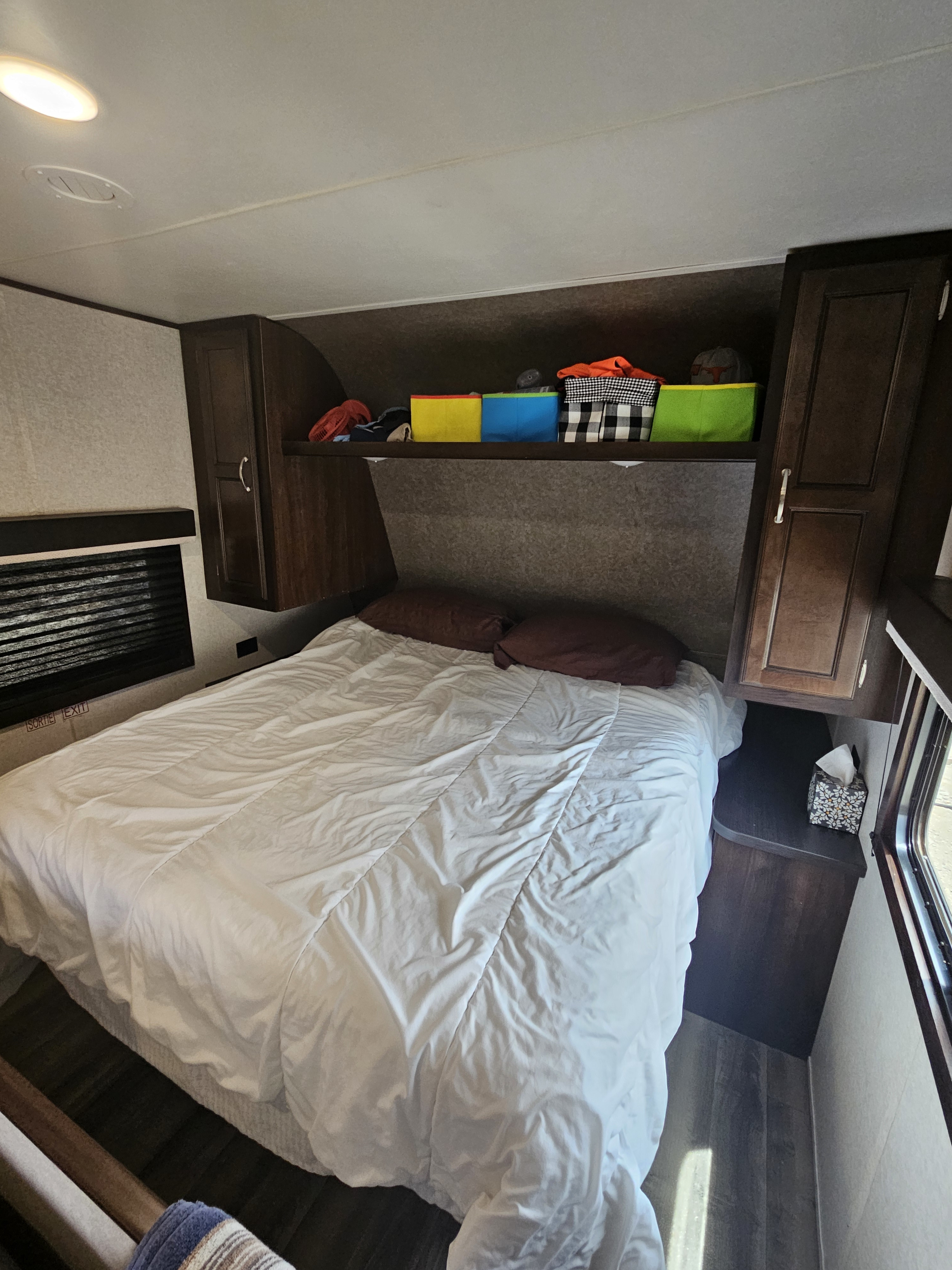 RV rental bedroom with a white-covered bed, dark wood cabinets, and an open shelf above the bed holding colorful storage bins.. Jayco Jay Flight SLX Rocky Mountain Edition 2018