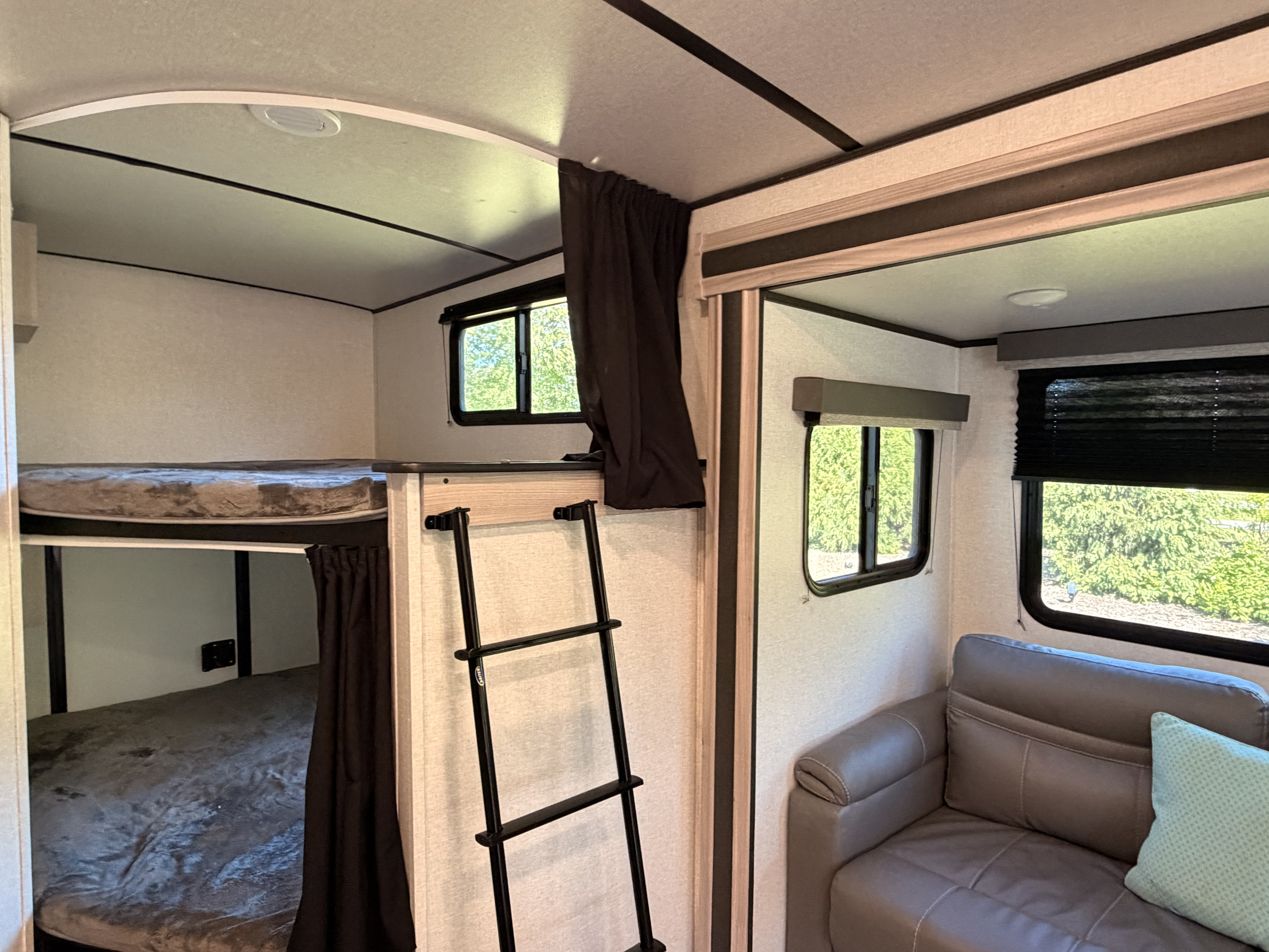 Interior of an RV rental: bunk beds with a ladder, grey sofa, and windows showing green trees outside.. Coachmen 2963BH 2023