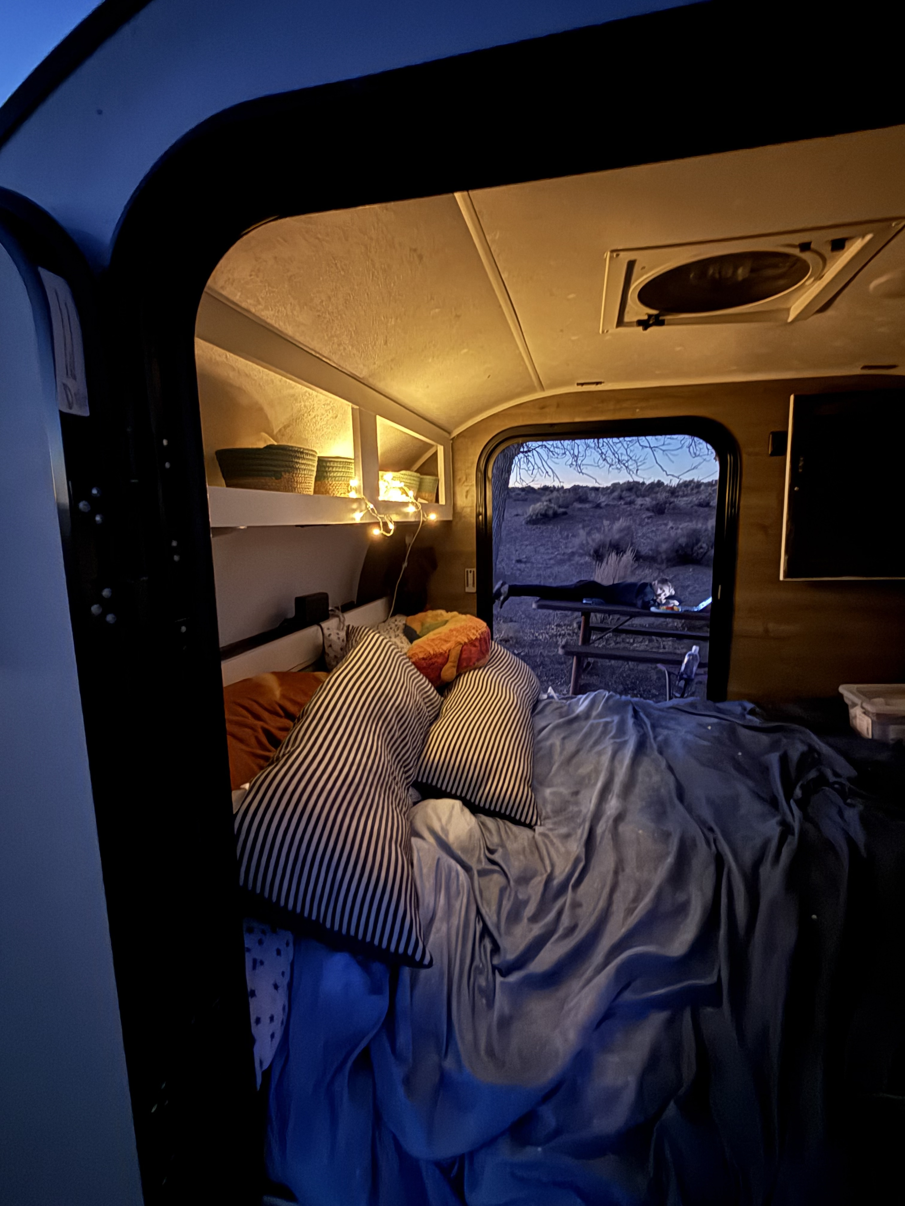 Cozy rv rental interior at dusk with a comfortable bed, string lights, and a person relaxing outside. Perfect for camping.. Braxton Creek Free Solo 2021