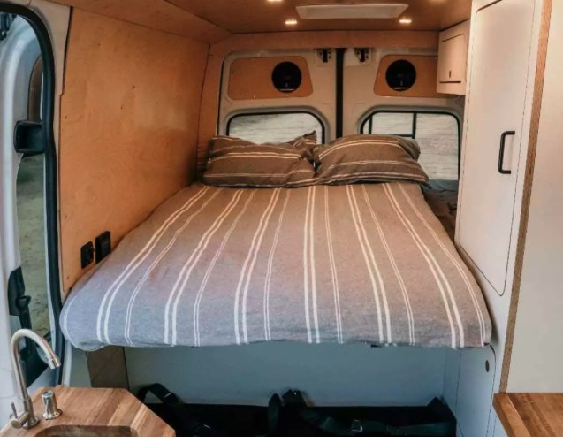 This RV rental features a comfy bed with striped bedding, wood walls, and a mini-kitchen sink, perfect for your camper van adventures.. Mercedes-Benz Sprinter 2023