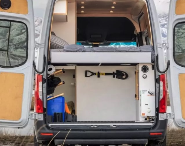 This rv rental shows a camper van's rear interior with a raised bed, gear storage, shovel mount, and a utility unit.. Mercedes-Benz Sprinter 2023