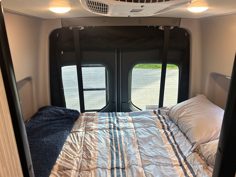 An RV rental's cozy camper van interior. A bed with striped comforter, rear windows, overhead lights & AC for comfortable travel.. Winnebago Revel 2023