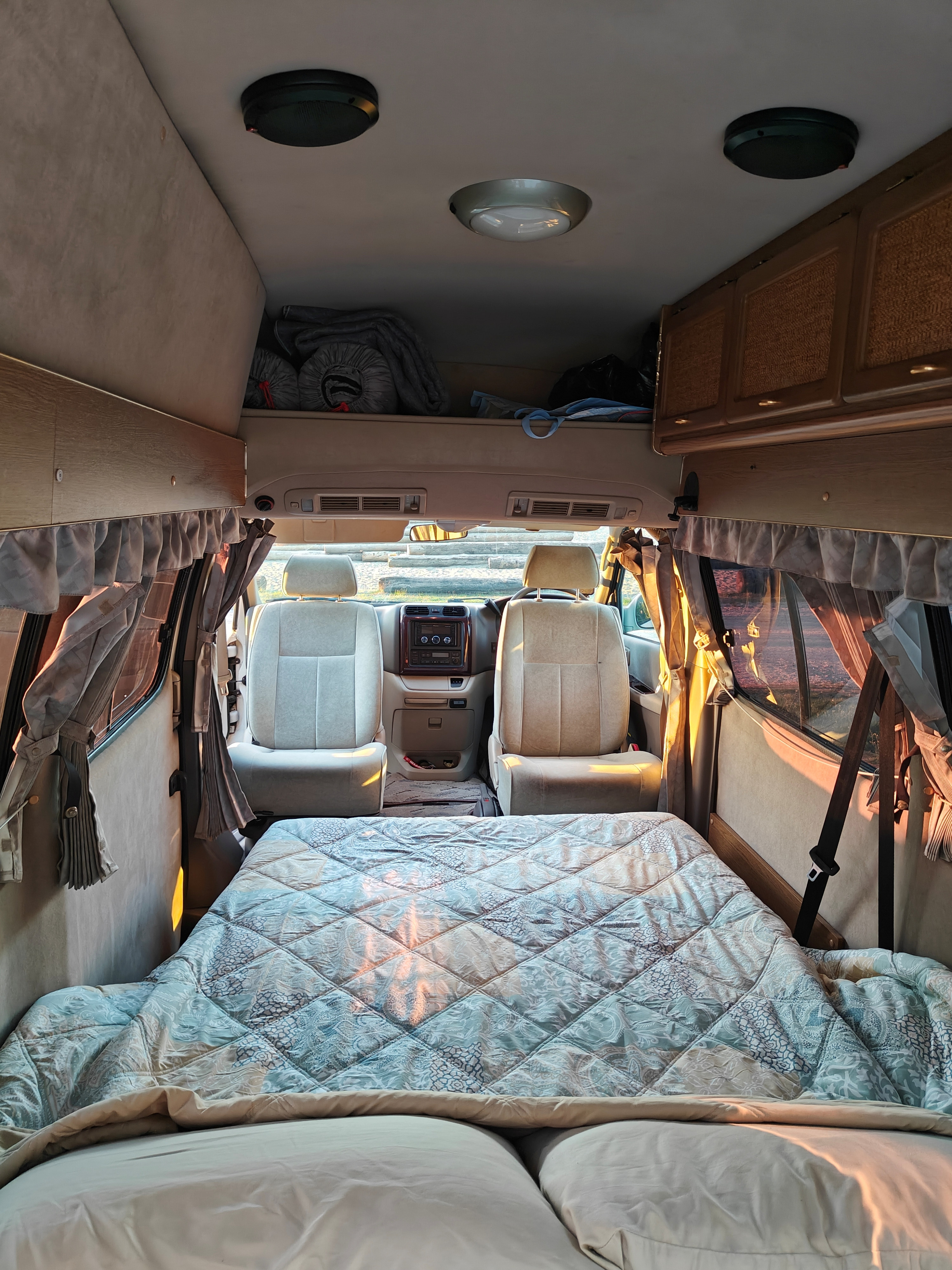 Comfortable rv rental interior: a plush bed in foreground, front seats and storage towards the driver's cabin, sunlit.. Toyota Hiace 4WD 2004