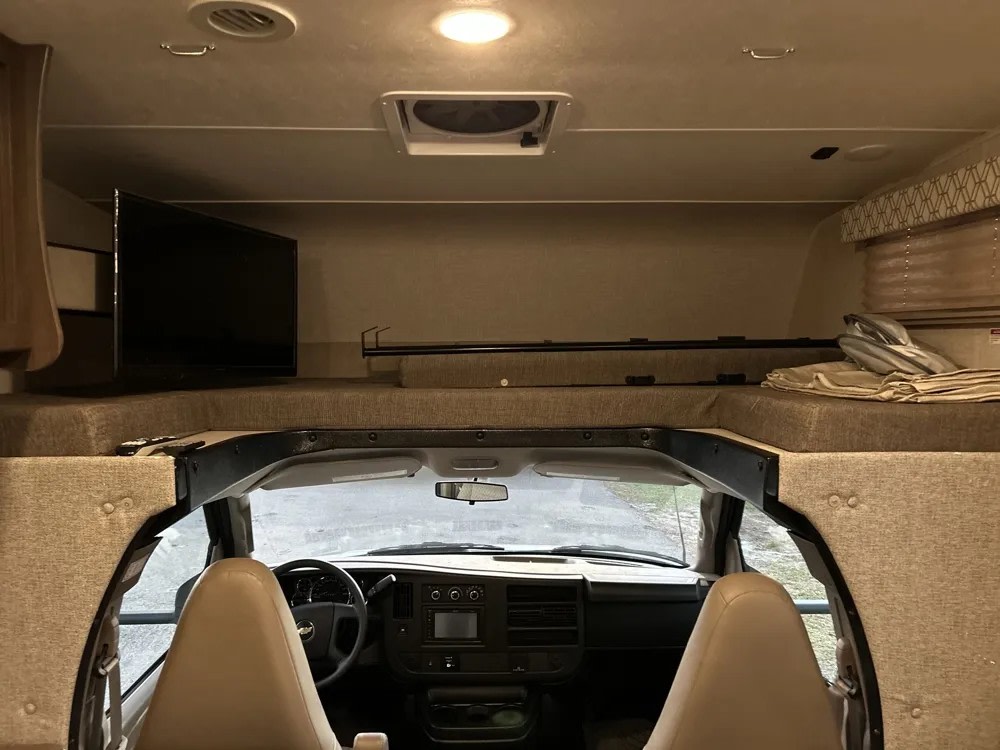 Cozy RV rental interior: cab-over bunk bed with TV & bedding, overlooking the driver's cockpit and dashboard.. Forest River Coachmen Freelander 2020