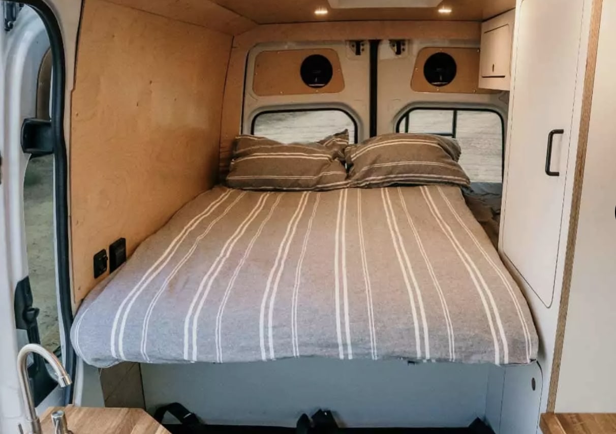 Cozy RV rental camper van interior. A neatly made bed with striped bedding fills the space, flanked by wood walls and a sink.. Mercedes-Benz Sprinter 2022