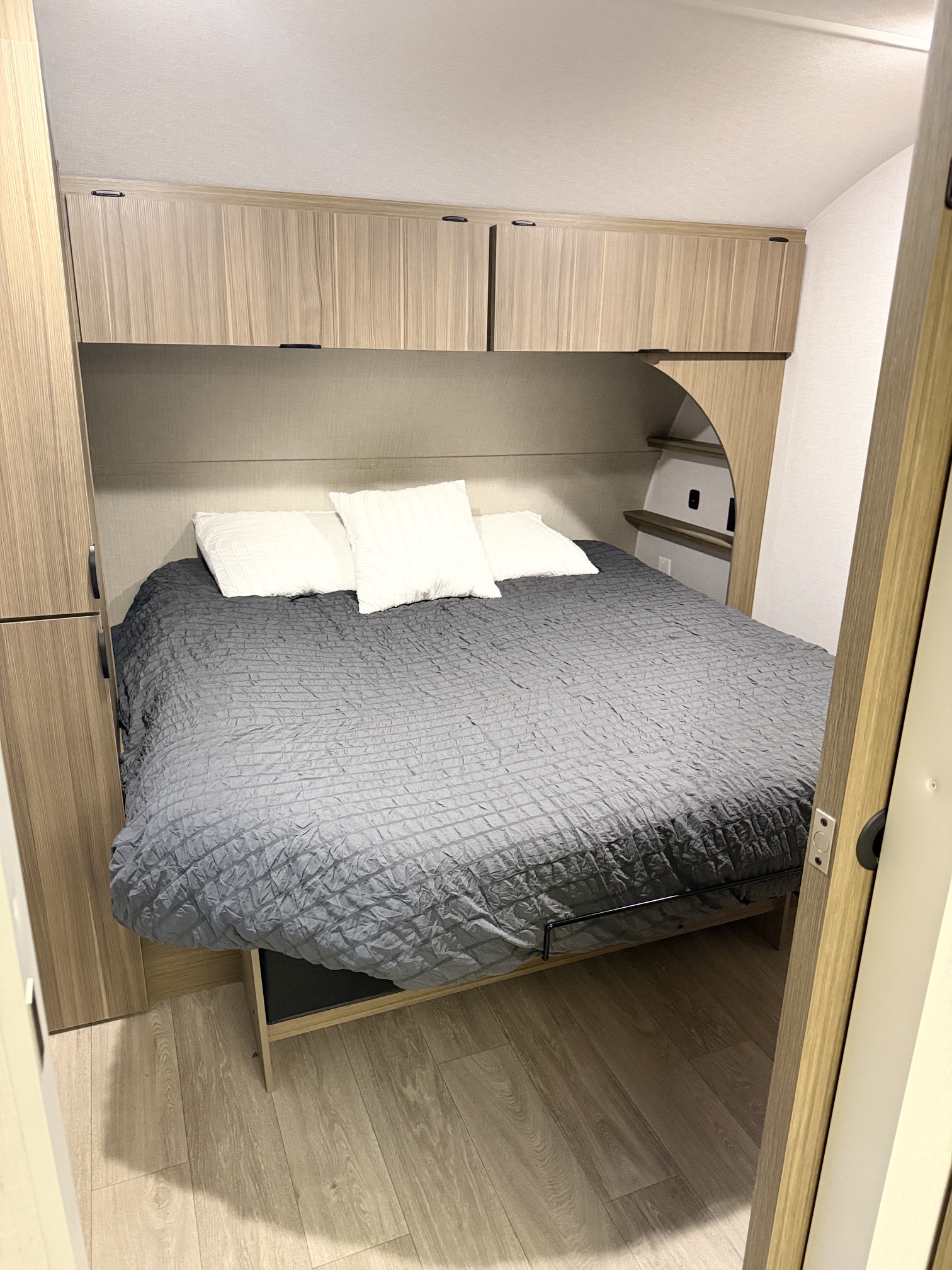 An RV rental bedroom features light wood cabinets, a grey quilted bed, white pillows, and side storage for a cozy space.. Forest River Wildwood X-Lite 2025