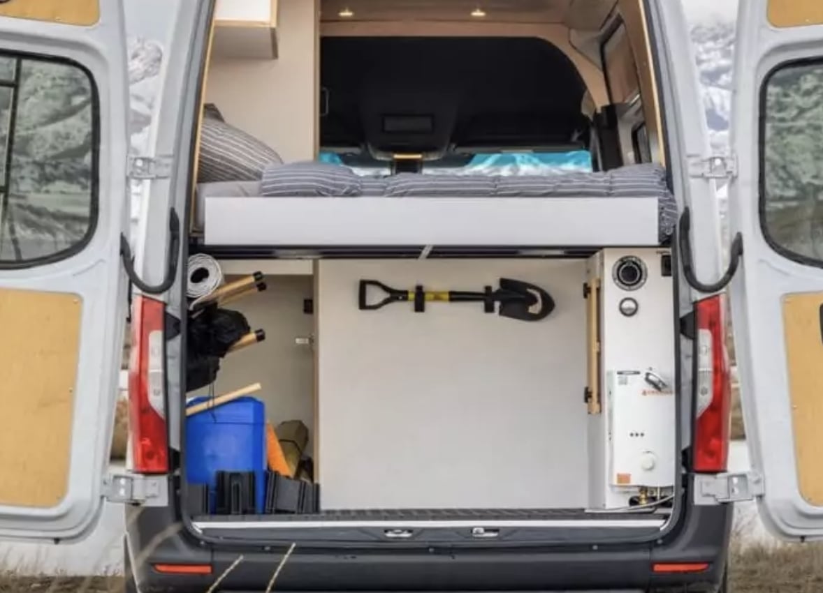 Inside an rv rental van: a raised bed, cleverly organized storage for camping gear, a mounted shovel, and a water heater.. Mercedes-Benz Sprinter 2024