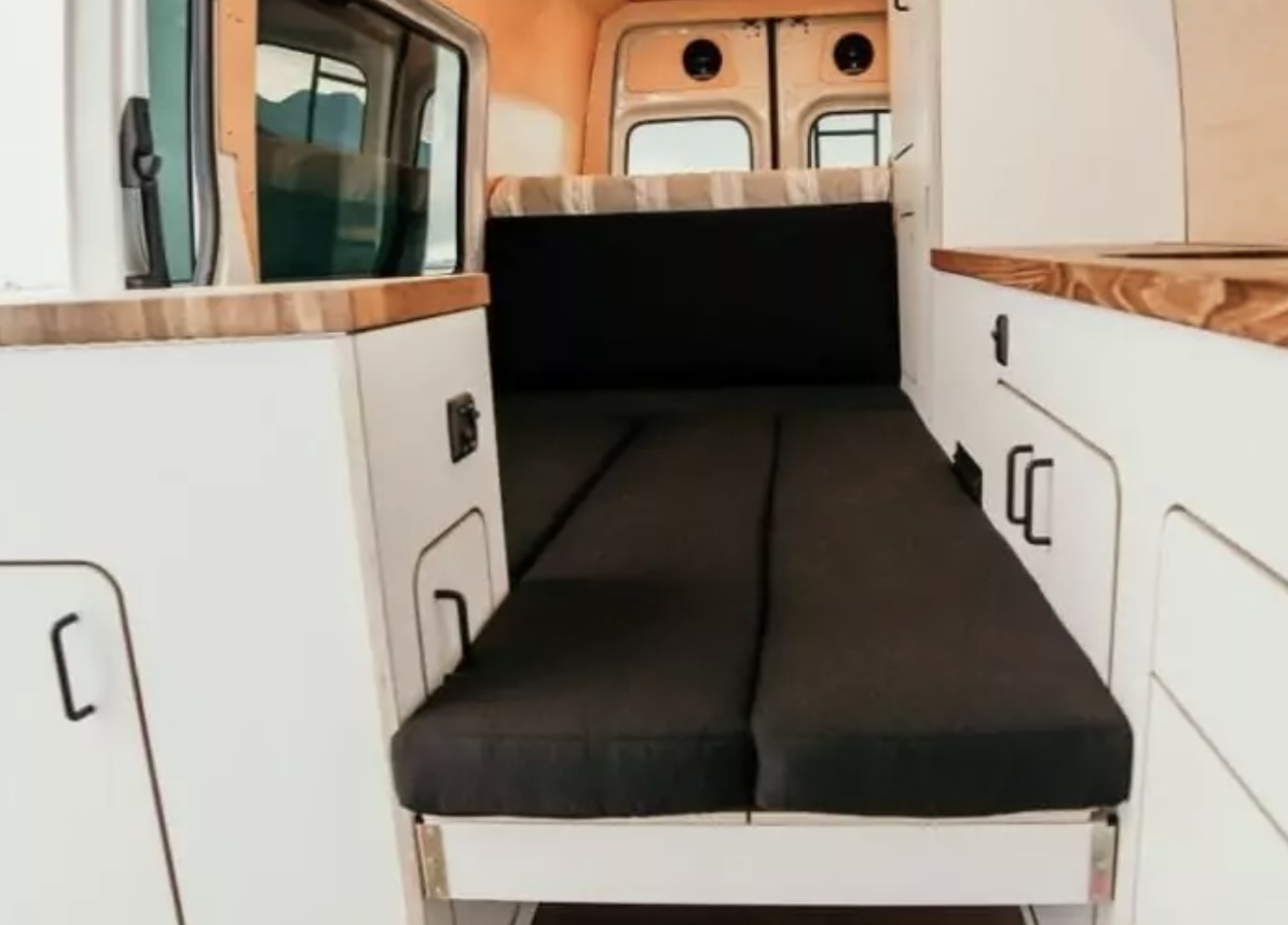 RV rental: Camper van interior shows a cozy black cushioned bed, white cabinets, and wood counters for comfy travel.. Mercedes-Benz Sprinter 2023