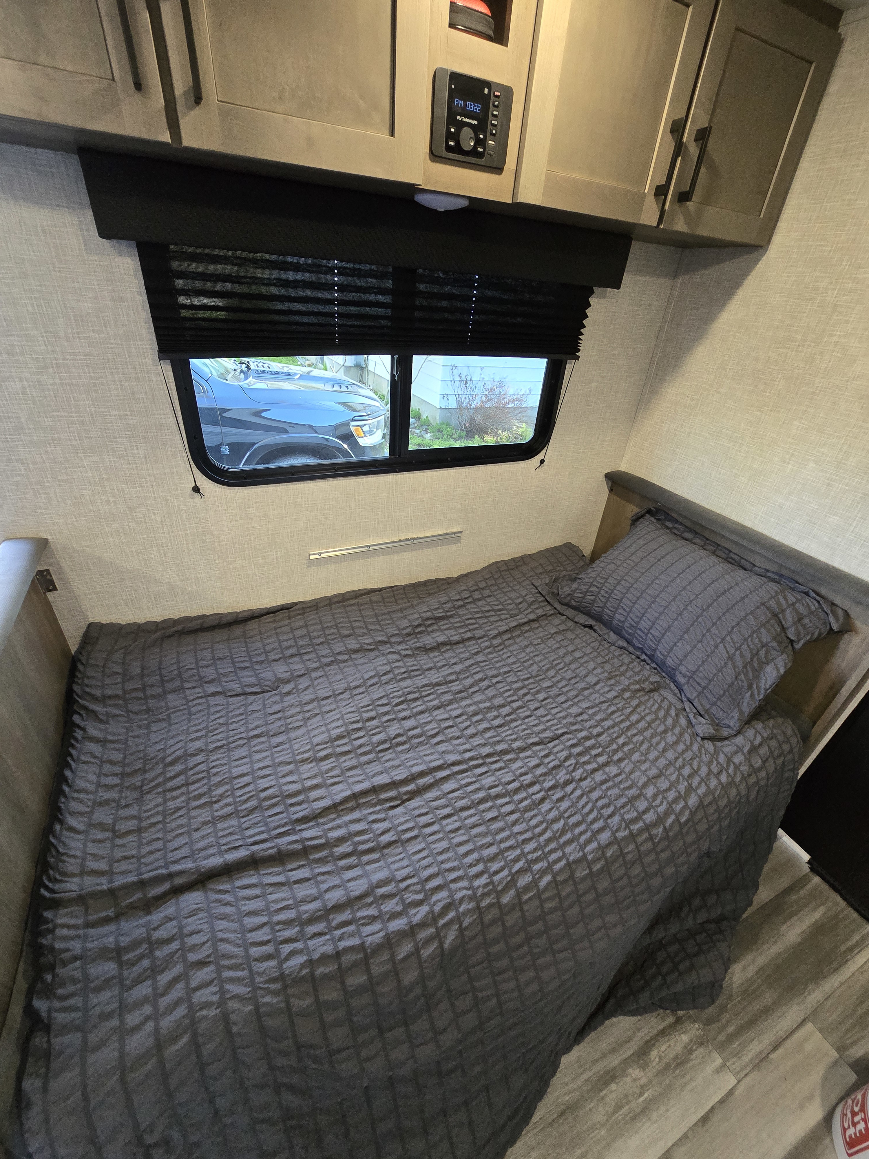 RV rental bedroom featuring a grey bed, overhead cabinets, and a window with a pleated blind. A control panel is visible.. Jayco Jay Flight SLX 2025