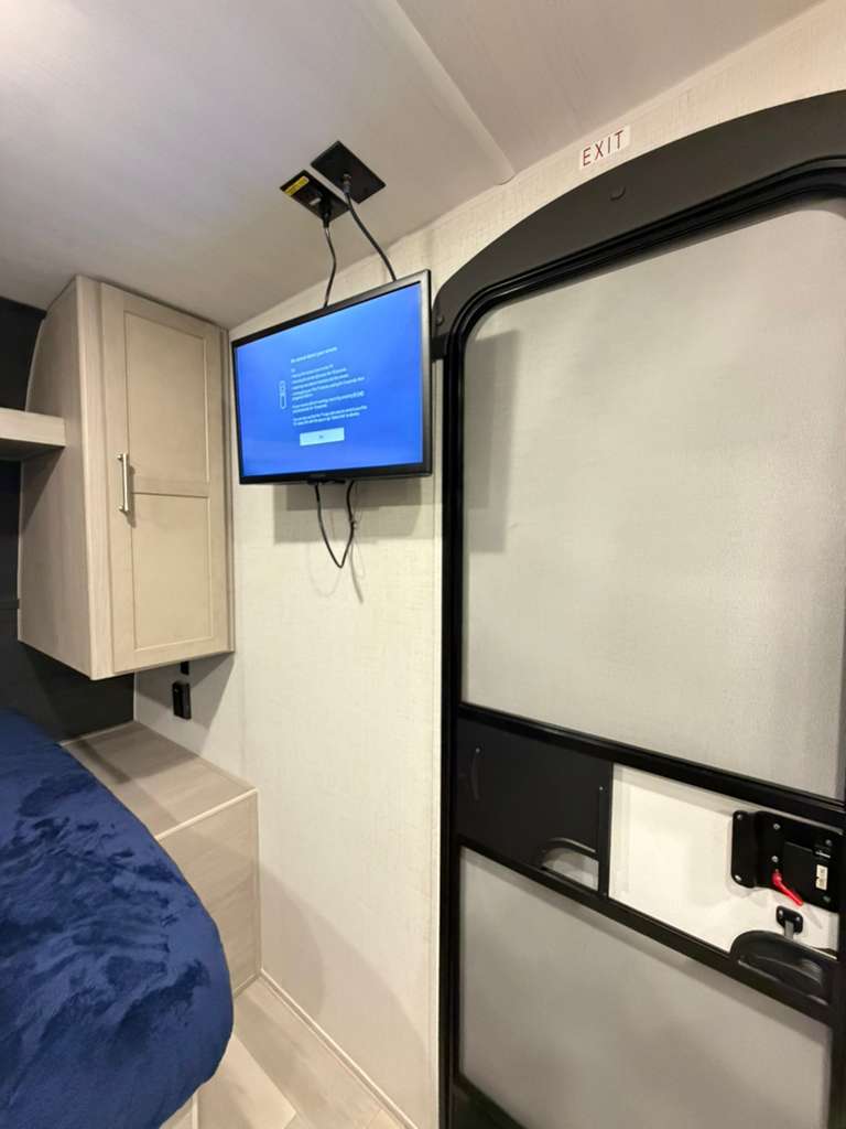 Cozy RV rental interior showing a TV, bed with blue blanket, light wood cabinet, and an exit door on the right.. Keystone RV Springdale 2024