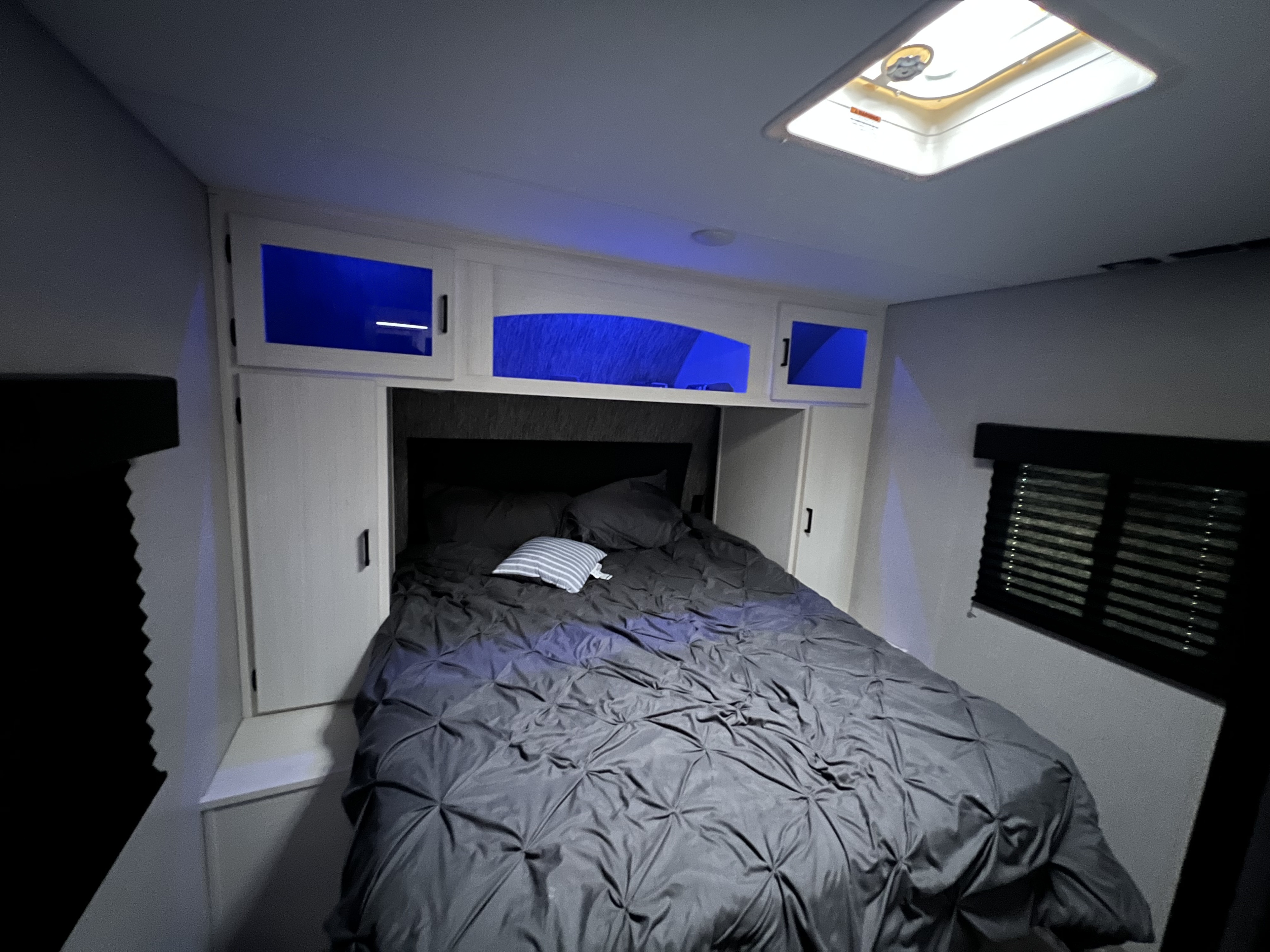 RV rental bedroom with dark gray bedding, blue lit overhead storage, and a bright skylight, offering a cozy compact space.. Venture Stratus 2021