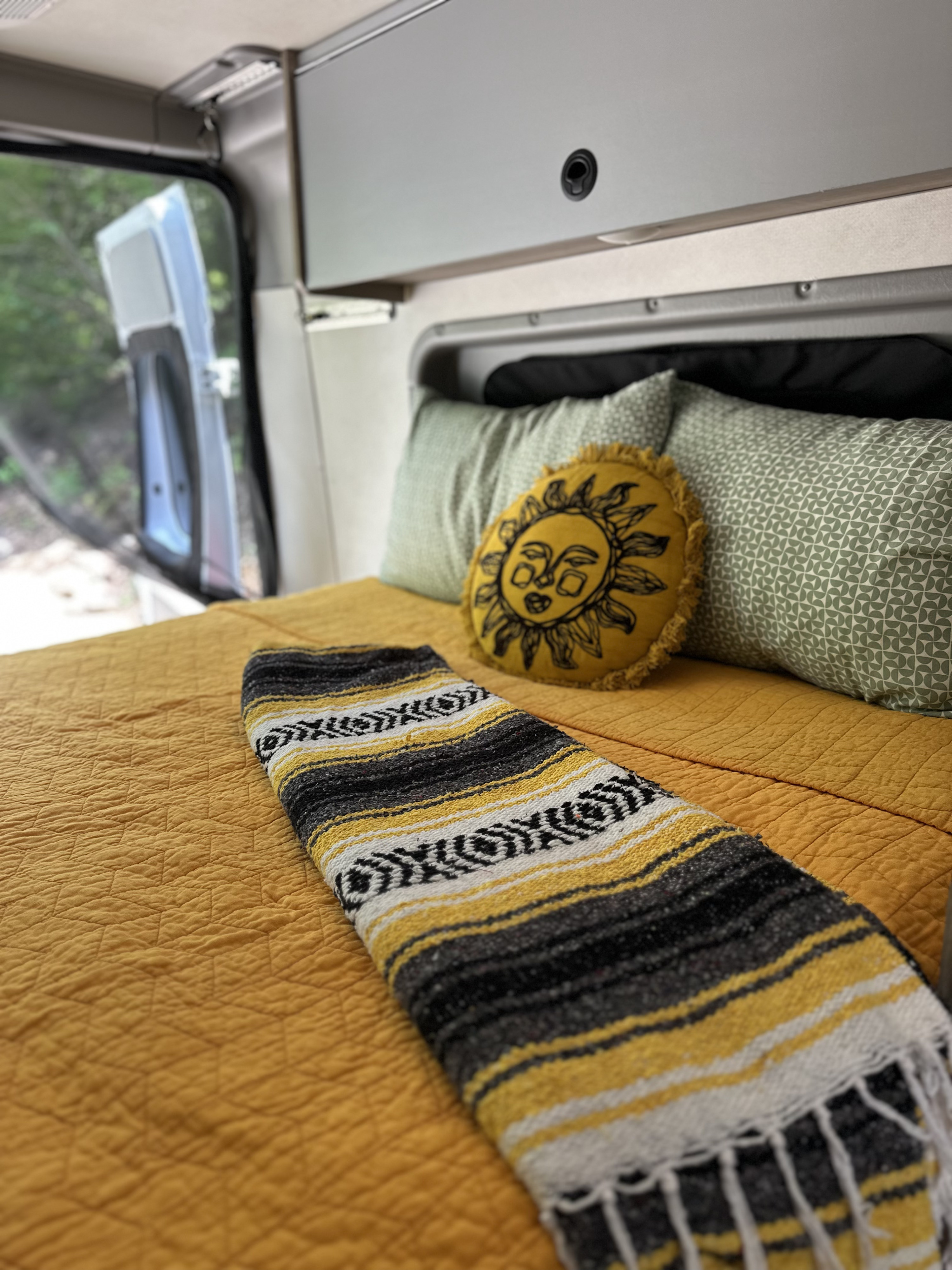Cozy RV rental interior featuring a yellow bed, round sun pillow, green patterned pillows, and a striped blanket, with nature view.. Winnebago Solis 59PX 2021