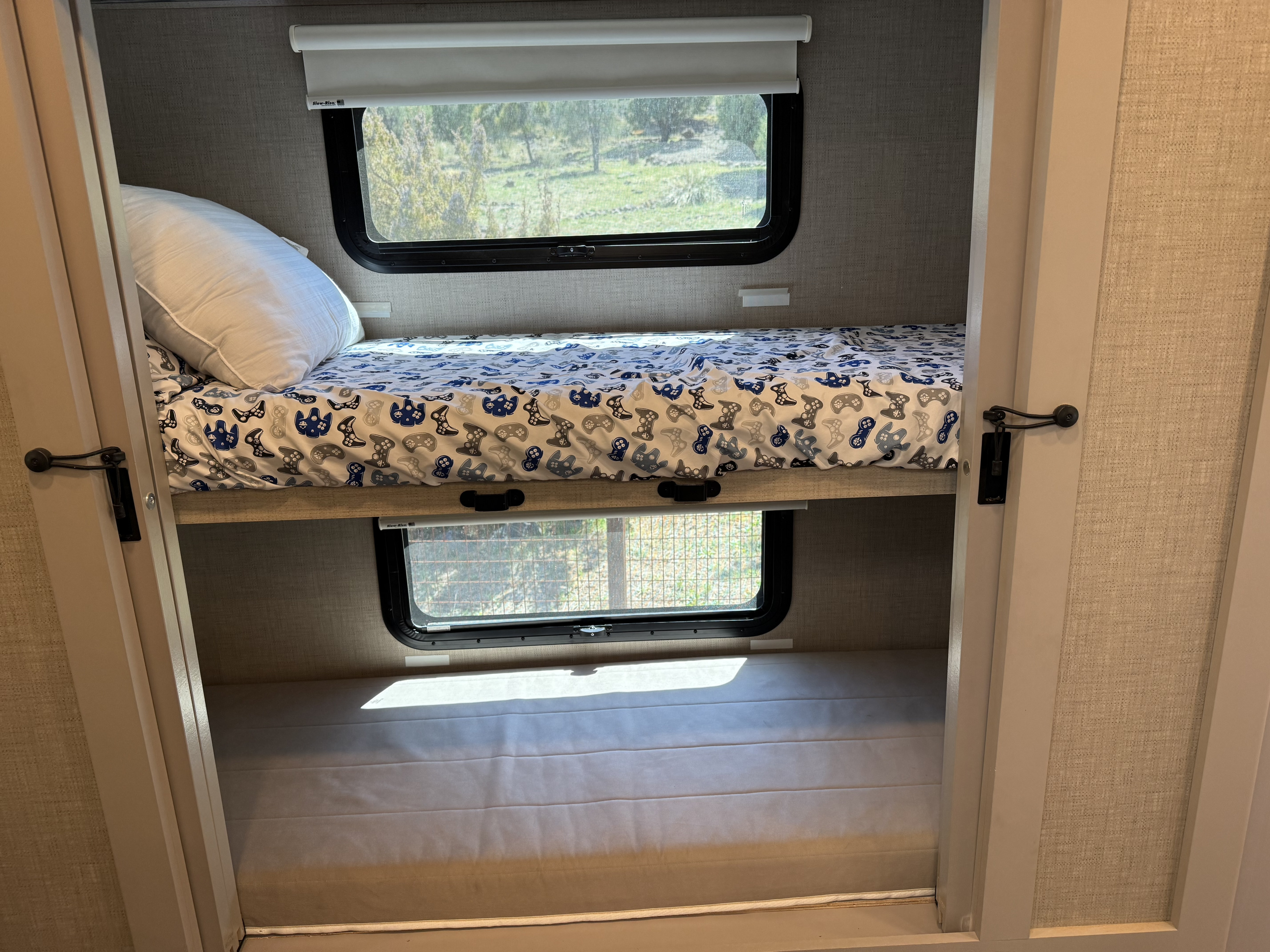 Cozy RV rental bunk beds with game controller sheets, bare mattress below, and windows showing an outdoor view.. Thor Magnitude 2023