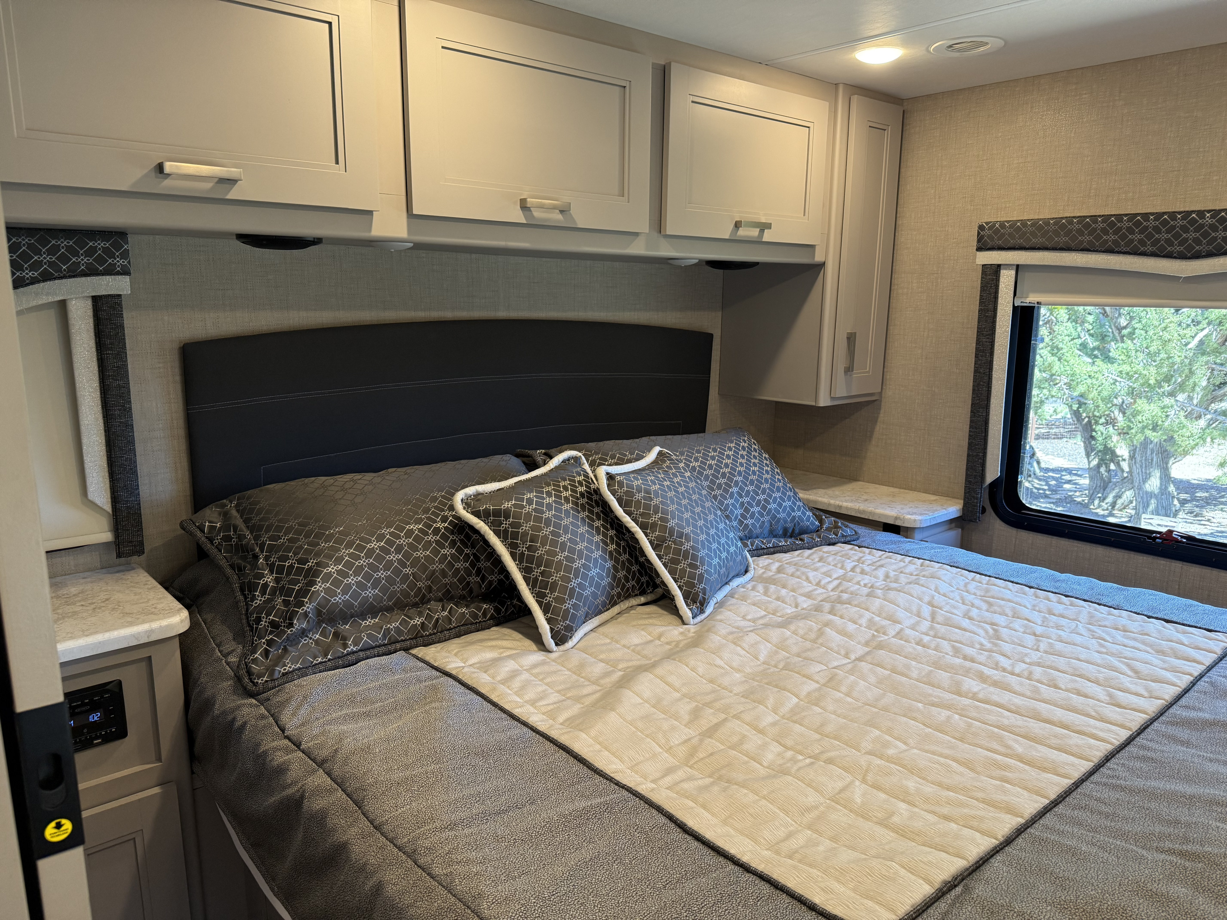 Comfortable RV rental bedroom featuring a bed with patterned pillows, overhead cabinets, and window with a tree view.. Thor Magnitude 2023