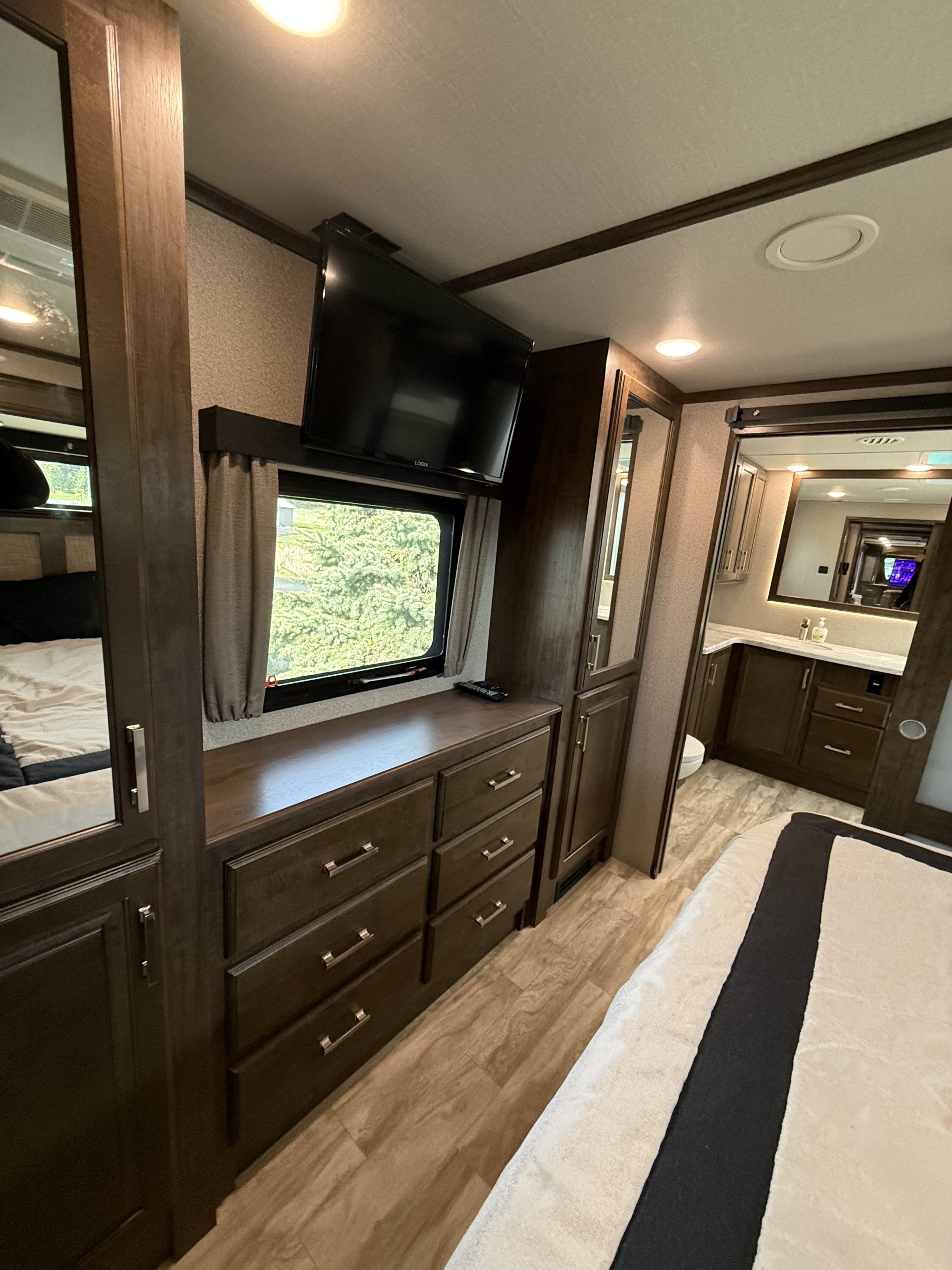 Cozy RV rental bedroom with dark wood dresser, mounted TV, window, and an en-suite bathroom visible beyond.. Grand Design Solitude 2020