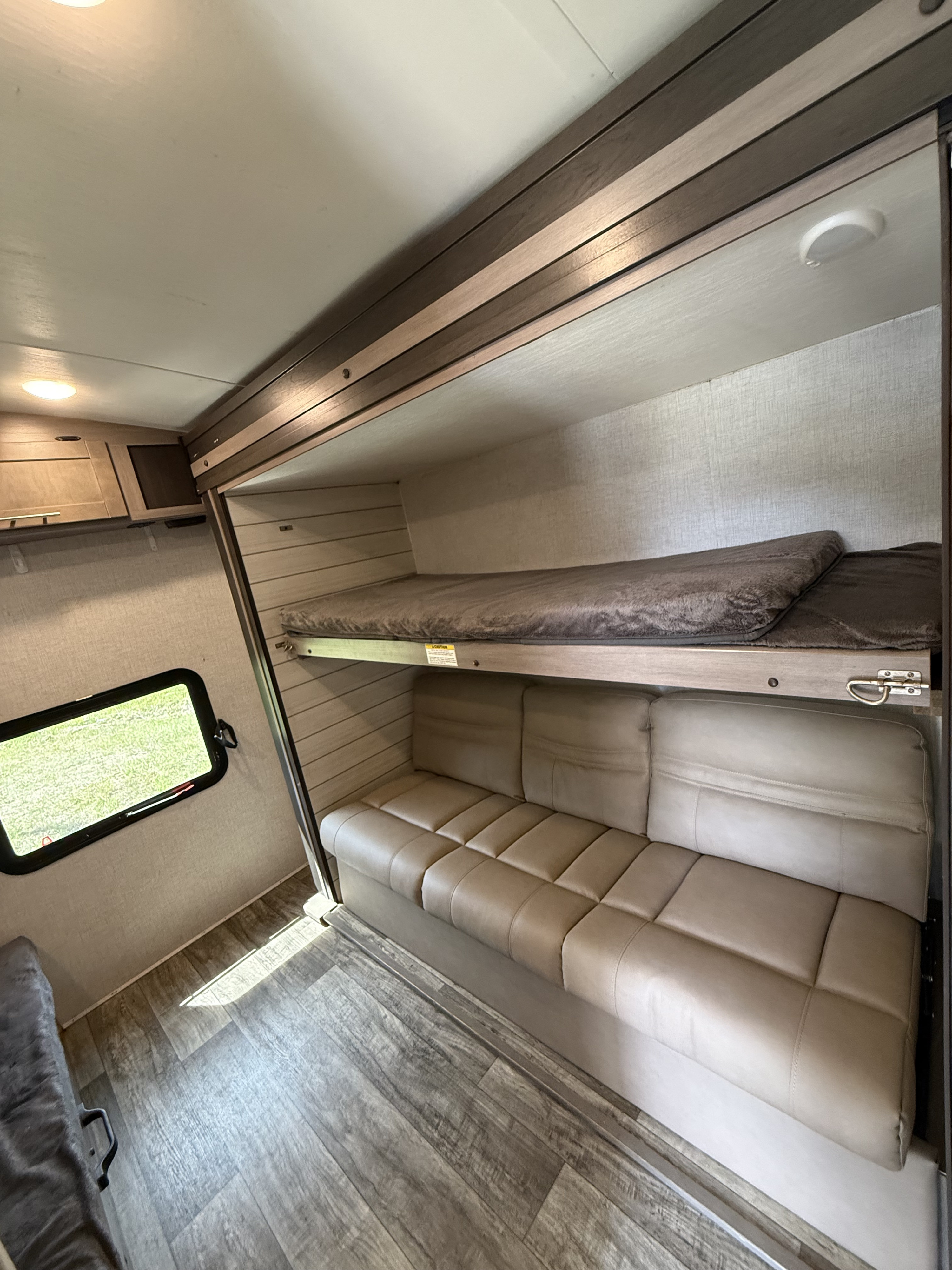 This RV rental interior shows a cozy bunk bed over a tan sofa, with a window view and wood-look flooring.. Crossroads Sunset Trail 36 ‘ 2022