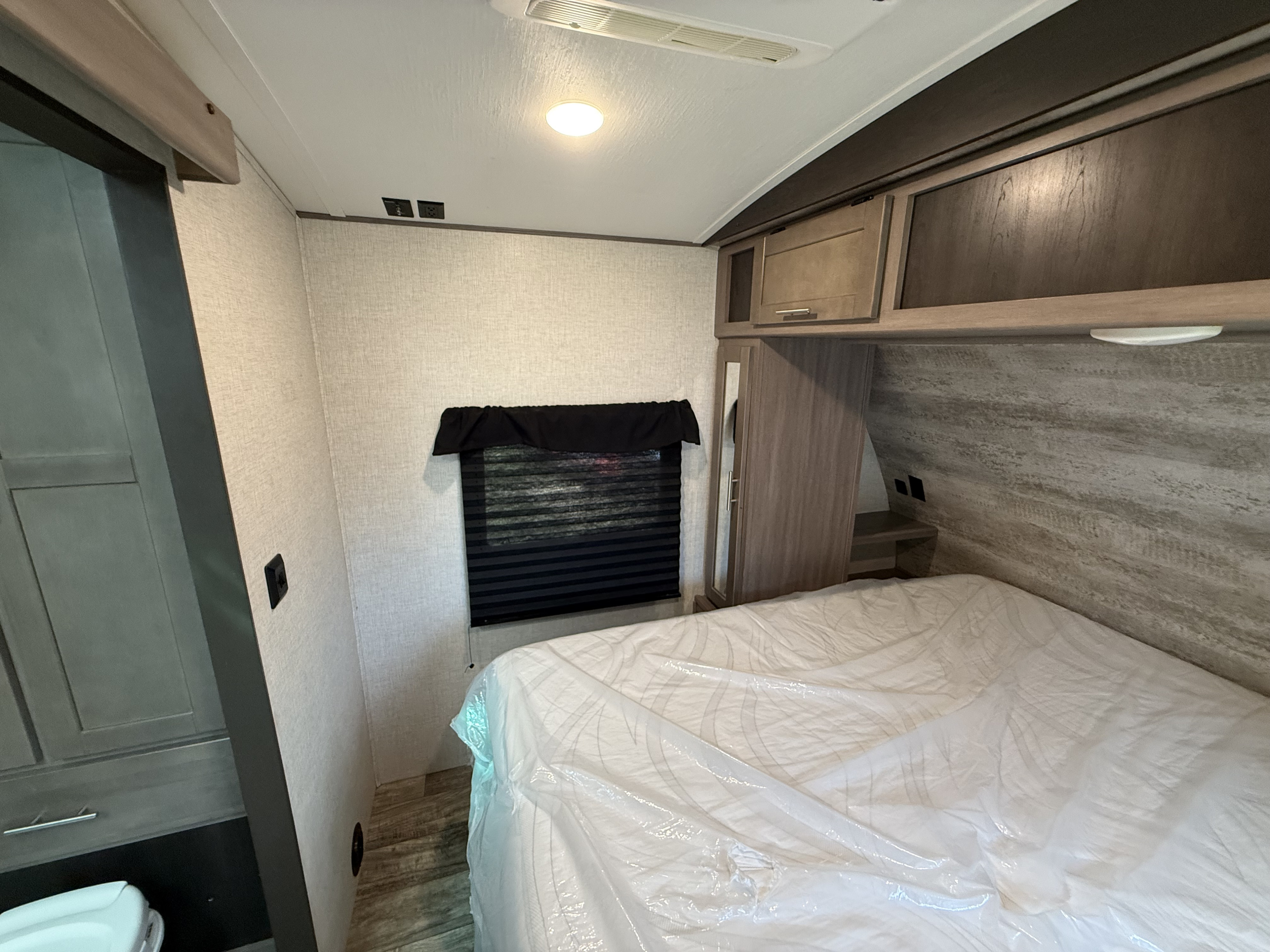 RV rental bedroom showcasing a bed with protective plastic, overhead cabinets, window, and wall-mounted outlets.. Crossroads Sunset Trail 36 ‘ 2022