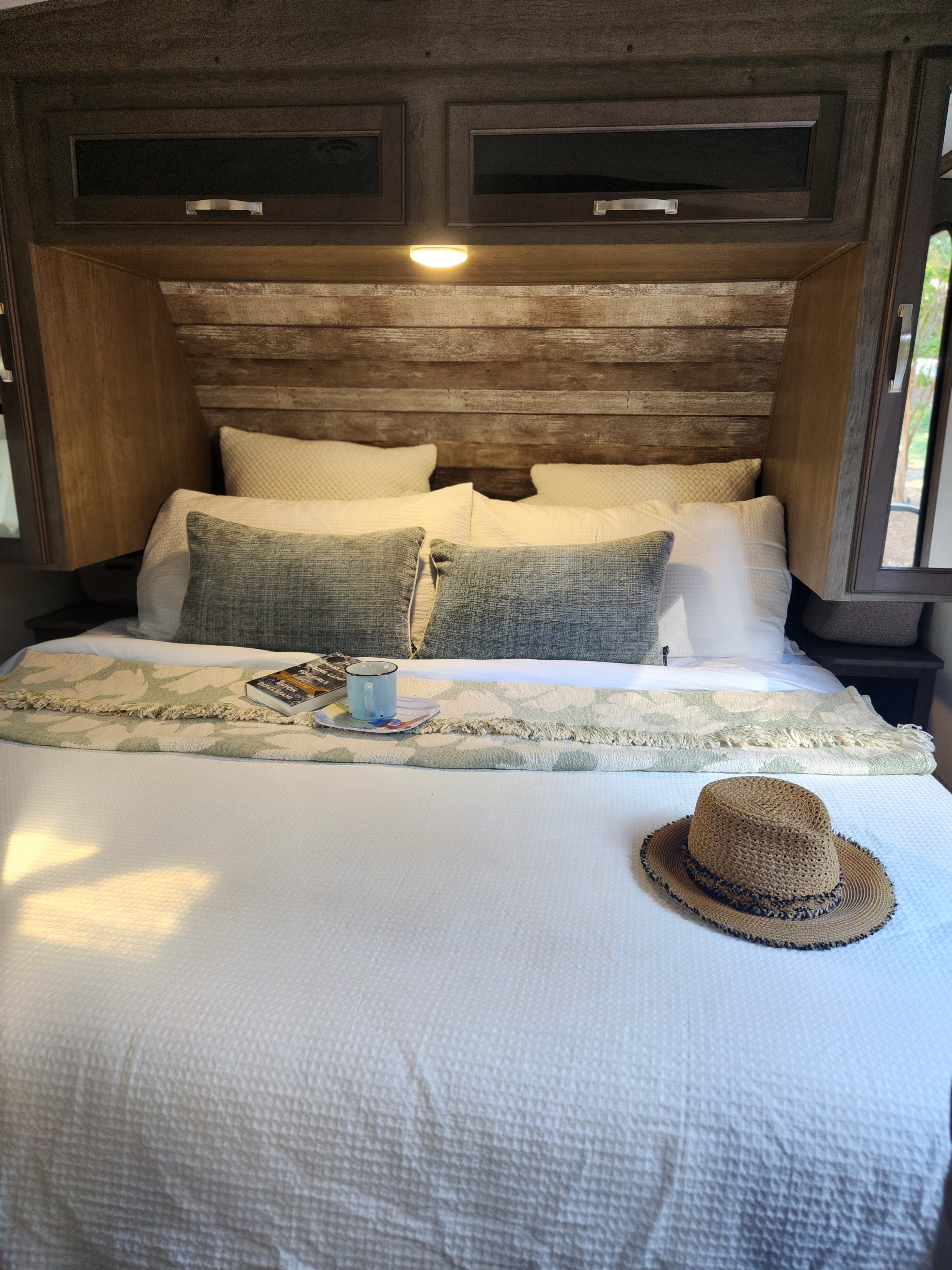 Cozy rv rental bedroom: A comfortable bed with pillows, a book, mug, and hat, illuminated by a warm overhead light.. Keystone RV Cougar 2023