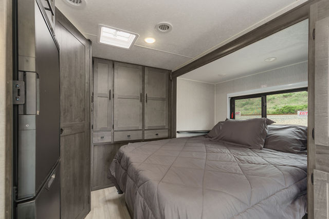 Cozy RV rental bedroom interior featuring a grey bed, built-in storage, window view, and overhead skylight.. Coachmen Prism 2022