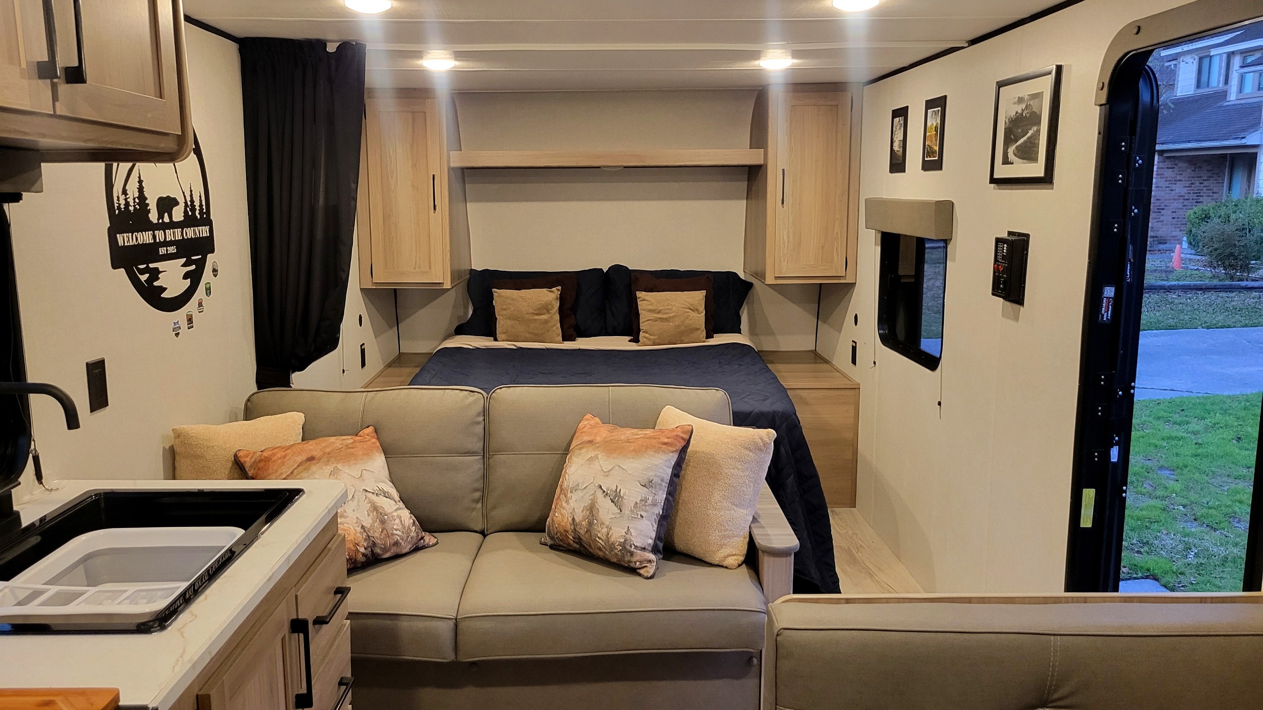 Inside a modern rv rental: a comfy bed, cozy sofa with mountain-themed pillows, and kitchen sink. Bright interior with outside view.. Dutchmen Coleman 2025