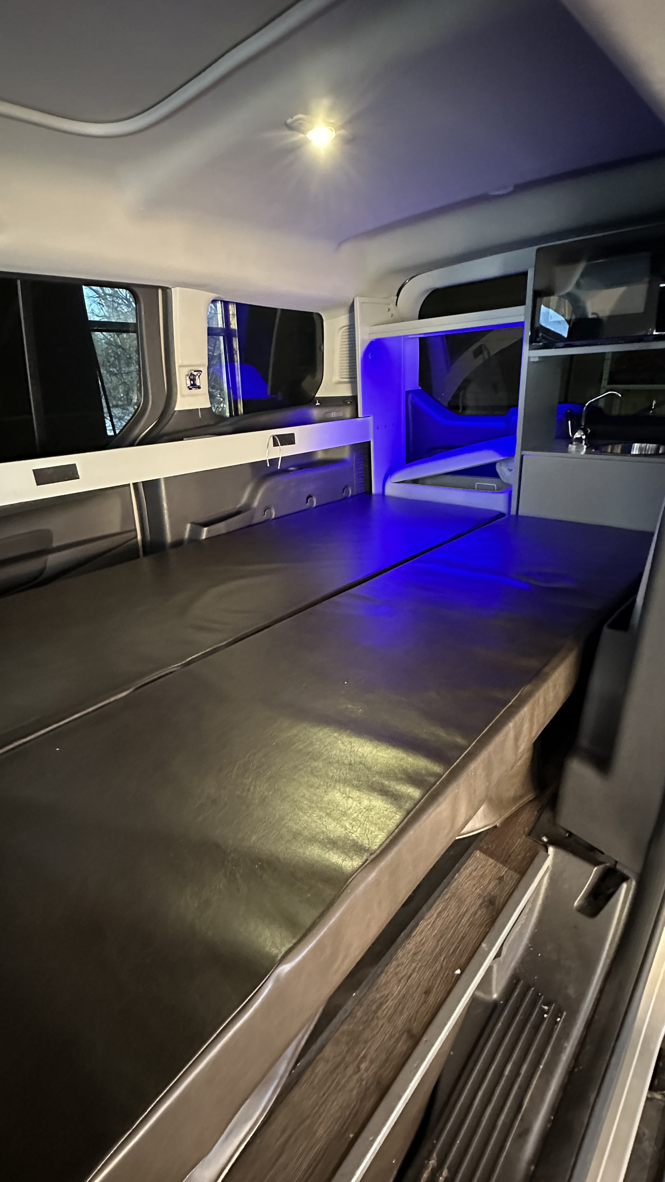 Inside an RV rental campervan: a cozy interior with a bed, windows, overhead lighting, and a compact kitchenette with blue light.. Ford Transit 2016