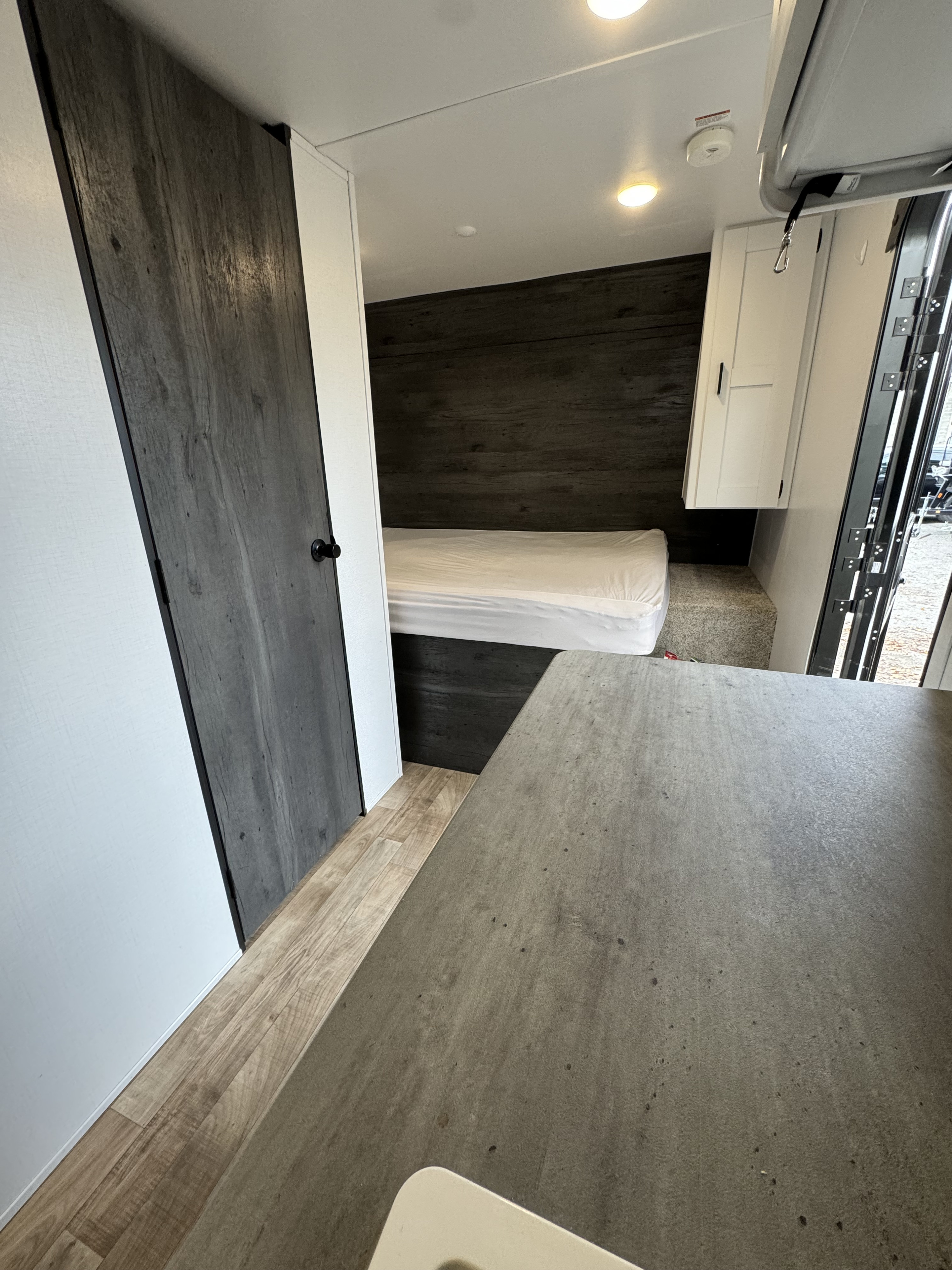 RV rental interior: A compact bedroom with a bed & dark accent wall. Light wood floor, gray counter in foreground, open side door.. Keystone RV Hideout 2022
