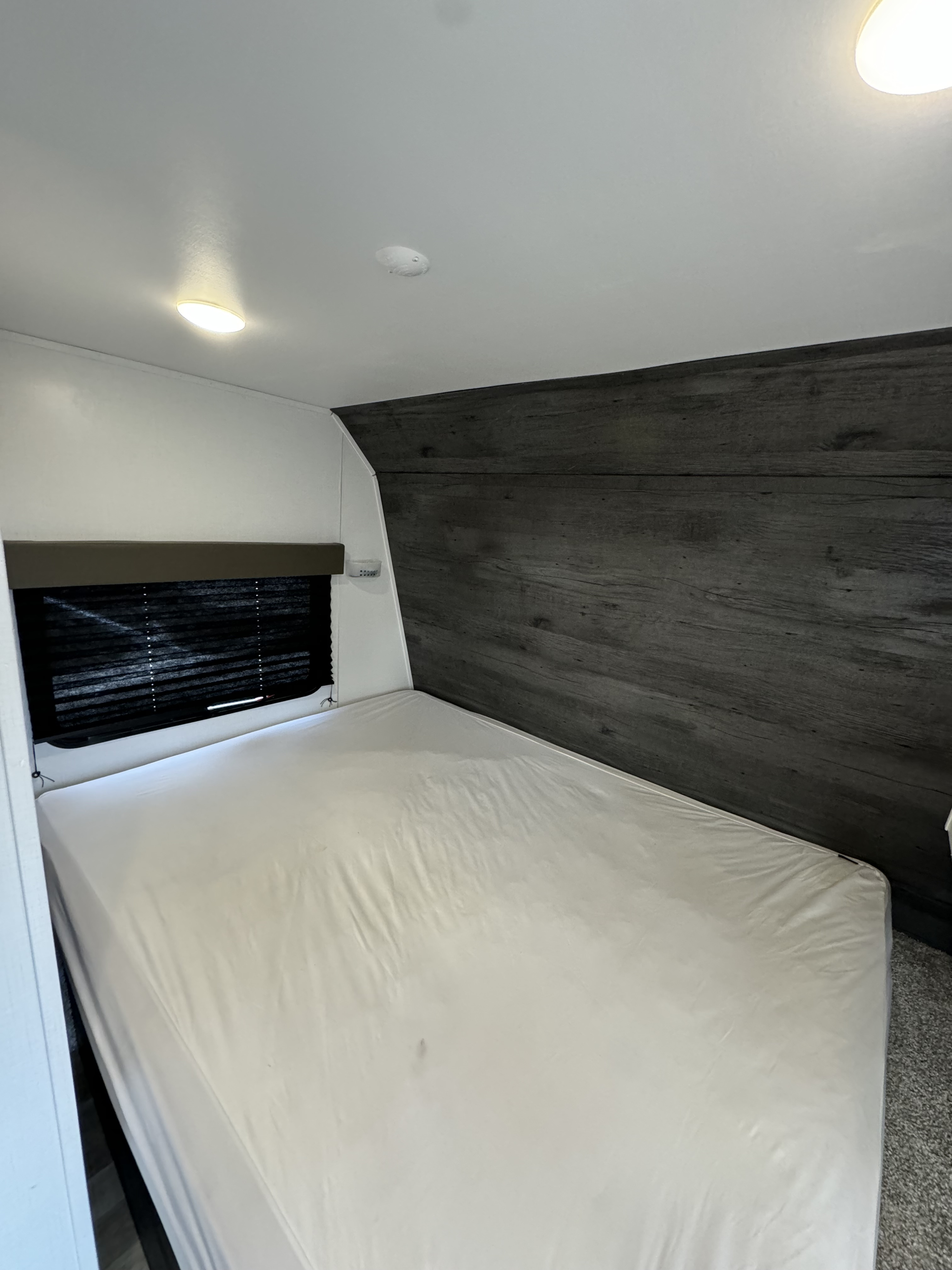 RV rental bedroom with a comfortable bed featuring white sheets, a dark window blind, and a modern dark wood-paneled wall.. Keystone RV Hideout 2022
