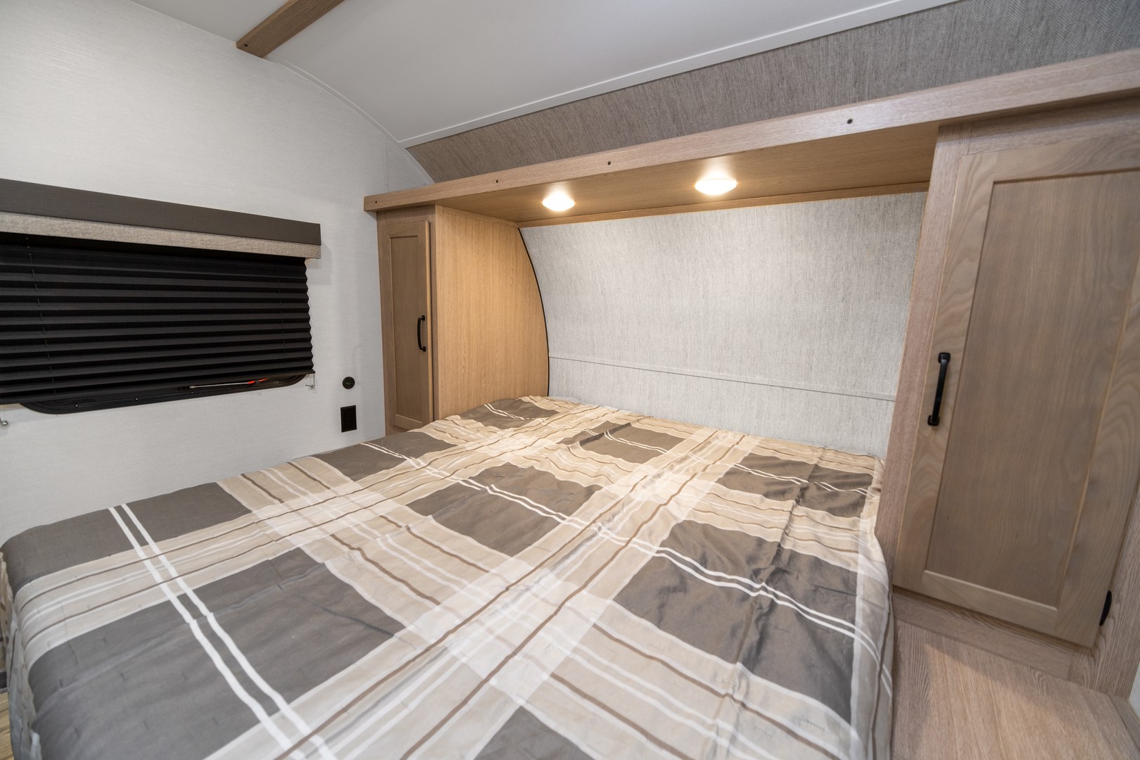RV rental bedroom showing a plaid bed, window with dark blinds, and built-in wood cabinetry with overhead lighting.. Gulf Stream Ameri-Lite 2021