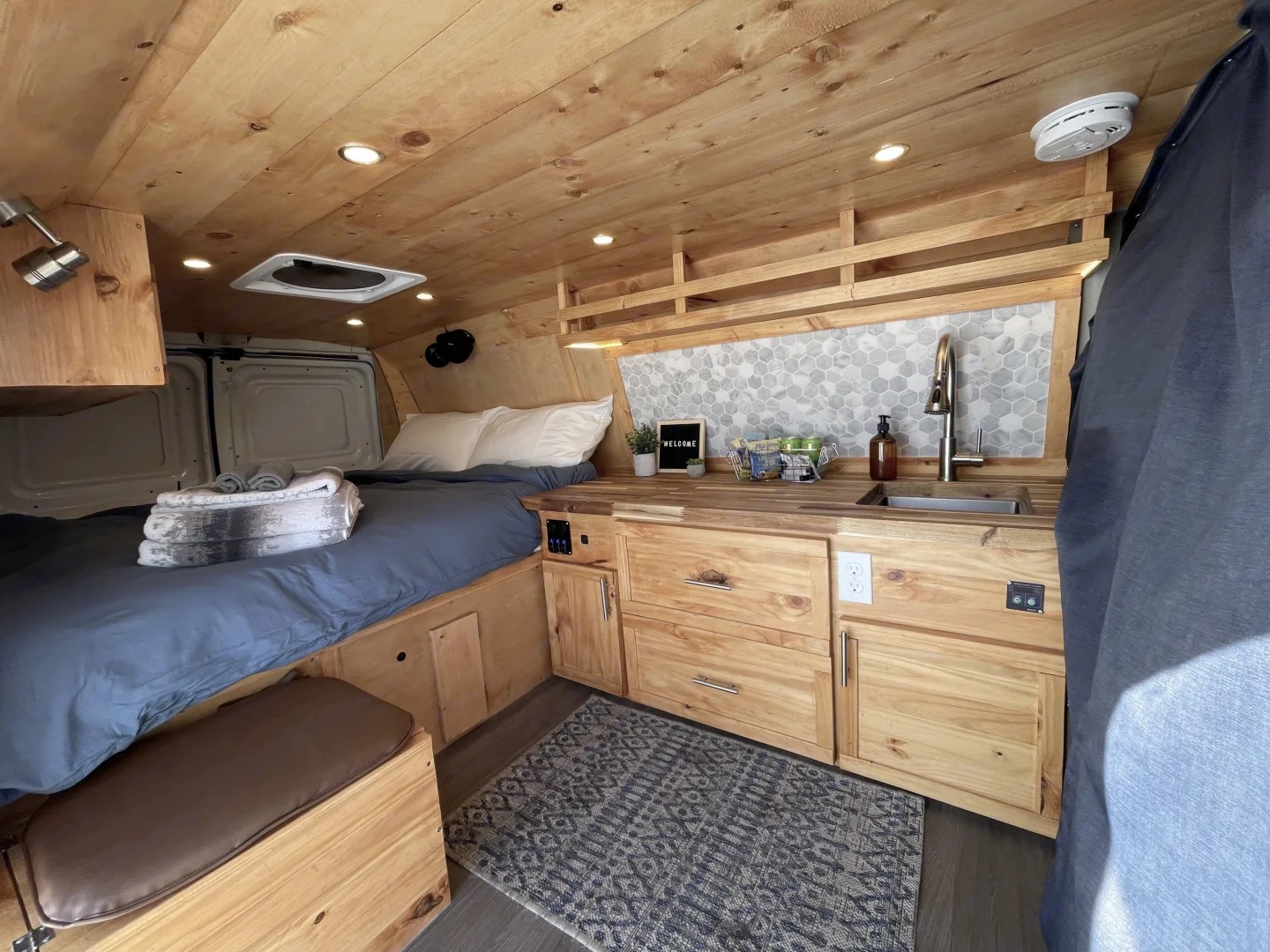 Inviting RV rental interior features a cozy bed, wooden kitchenette with sink, tile backsplash, and ample storage, all in a compact van.. Gmc Savanna 2500 2016