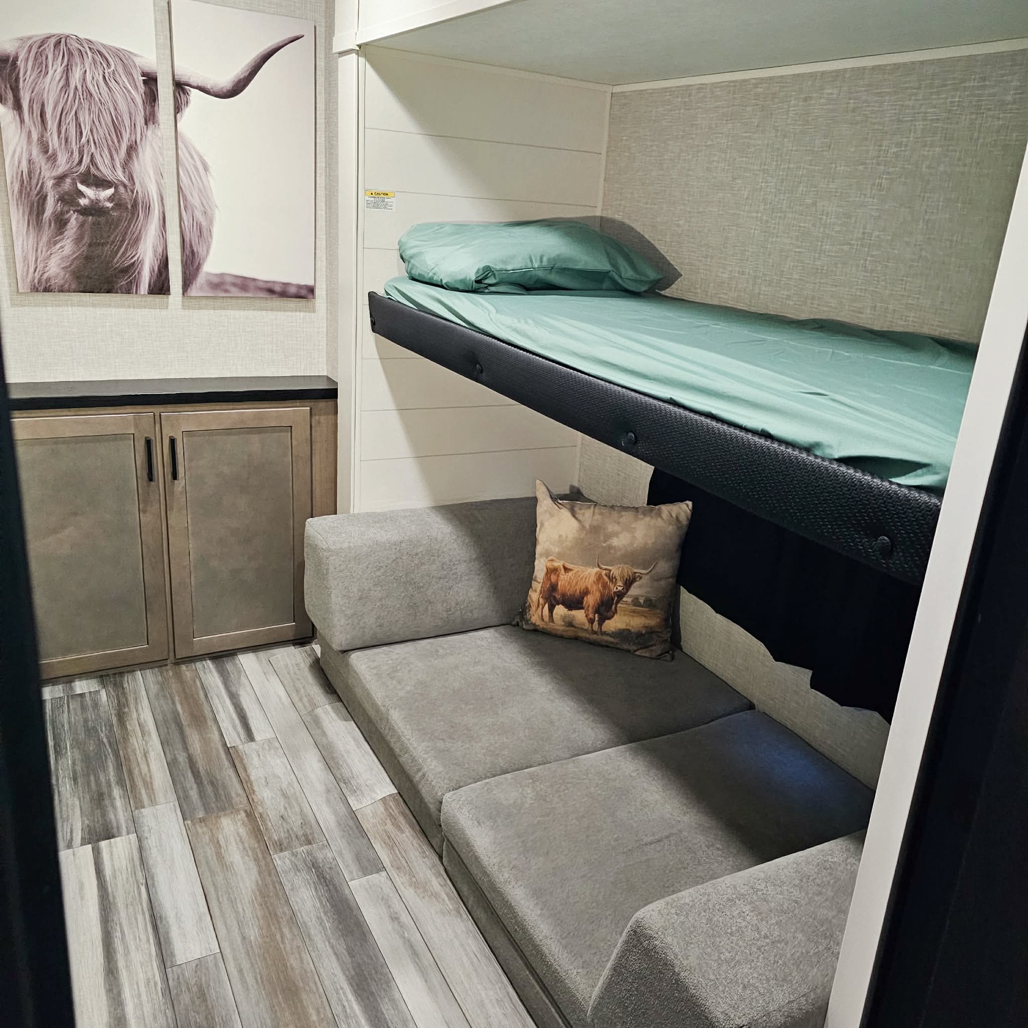 RV rental bunk bed setup with a gray sofa, Highland cow wall art, and cabinets. Green sheets on the bed.. Jayco SLX 2025