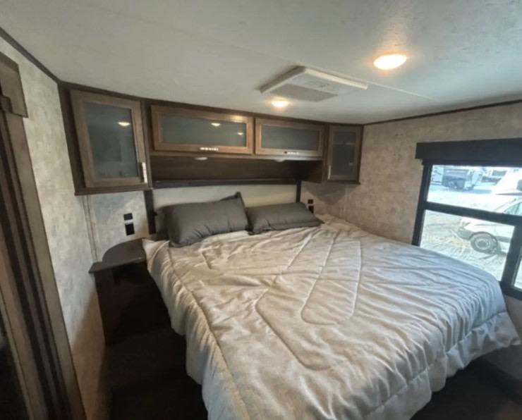 Cozy RV rental bedroom featuring a large bed with gray pillows, overhead cabinets, and a window view.. Highland Ridge RV Highlander Toy Hauler 2019