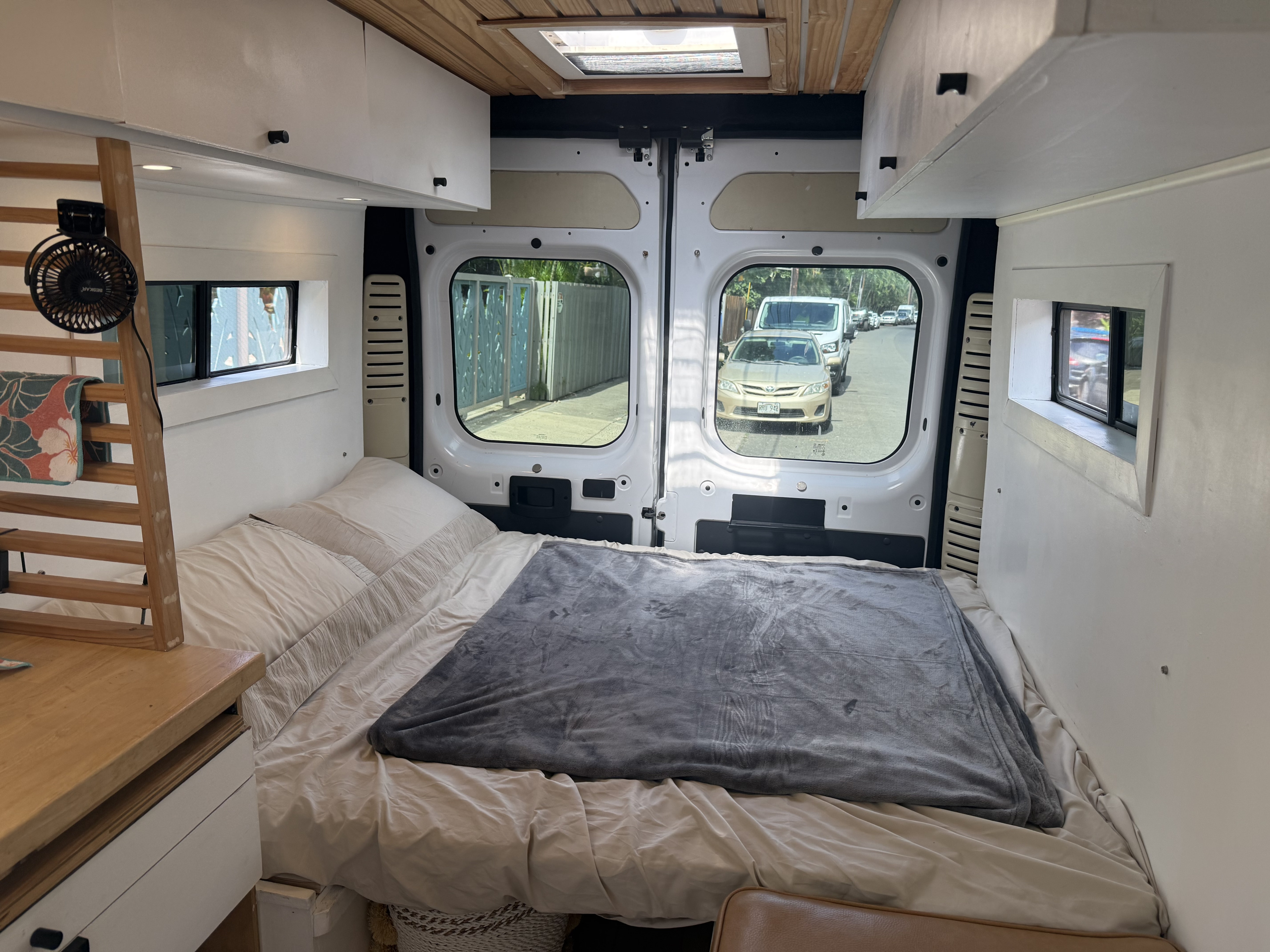 Cozy RV rental interior: a comfy bed, wooden shelves with a fan, and a view of the street through the open rear doors.. Ram ProMaster 2500 2019