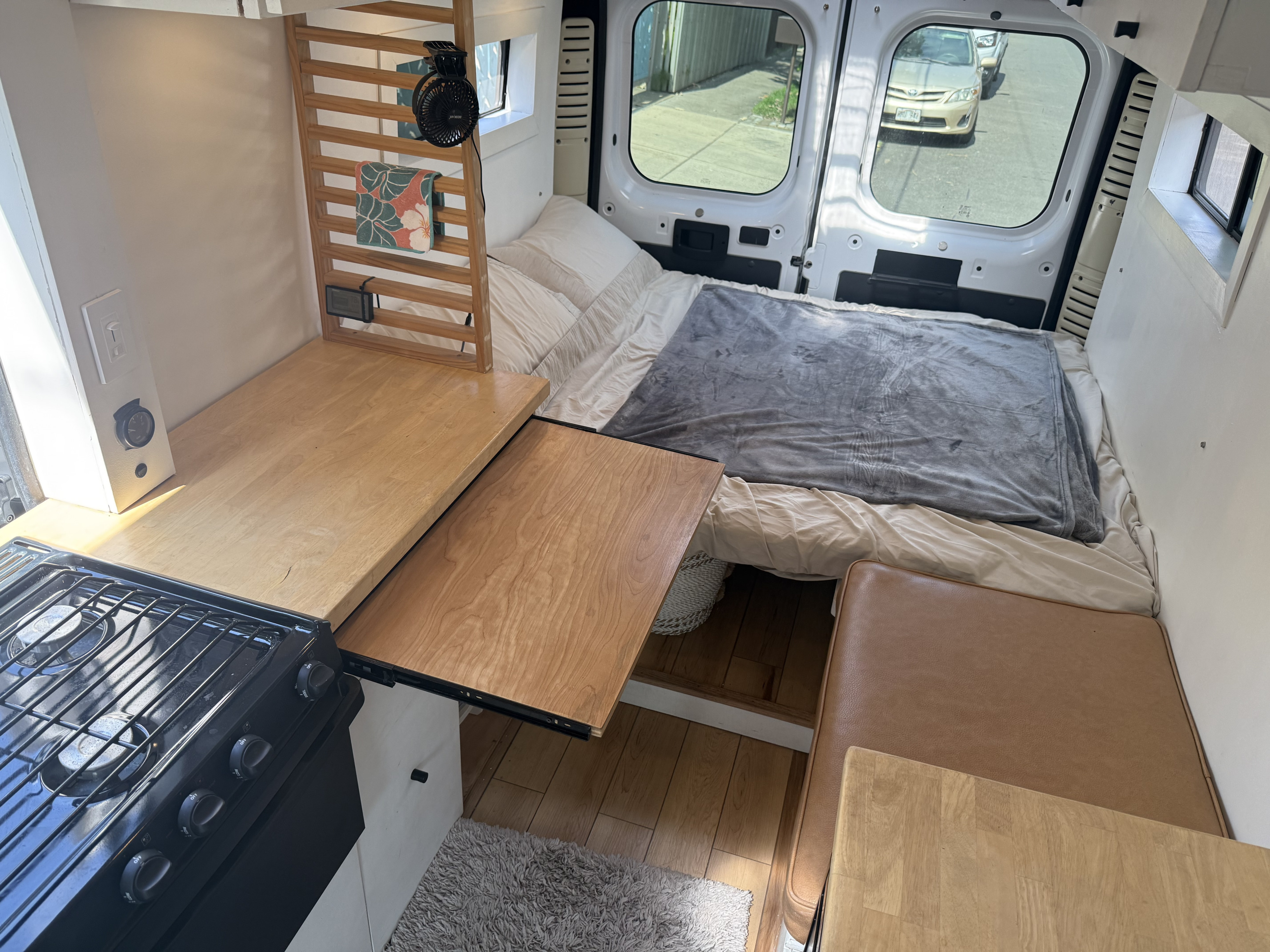 Inside an RV rental van, a full bed, gas stove with extending countertop, and seating create a compact living space.. Ram ProMaster 2500 2019