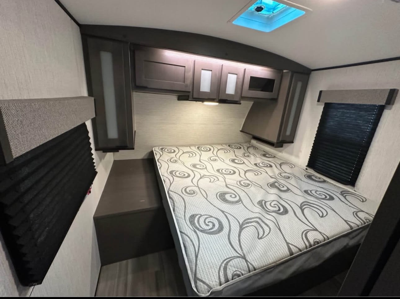 RV rental bedroom with a patterned bed, dark overhead cabinets, and blackout blinds. This compact interior is ready for travel.. Cruiser RV MPG 2022