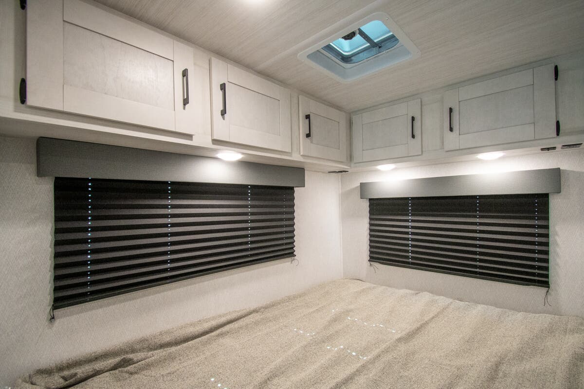 RV rental bedroom with light overhead cabinets, dark pleated blinds, and a neatly made bed, illuminated by ceiling lights.. Forest River Entrada 2026