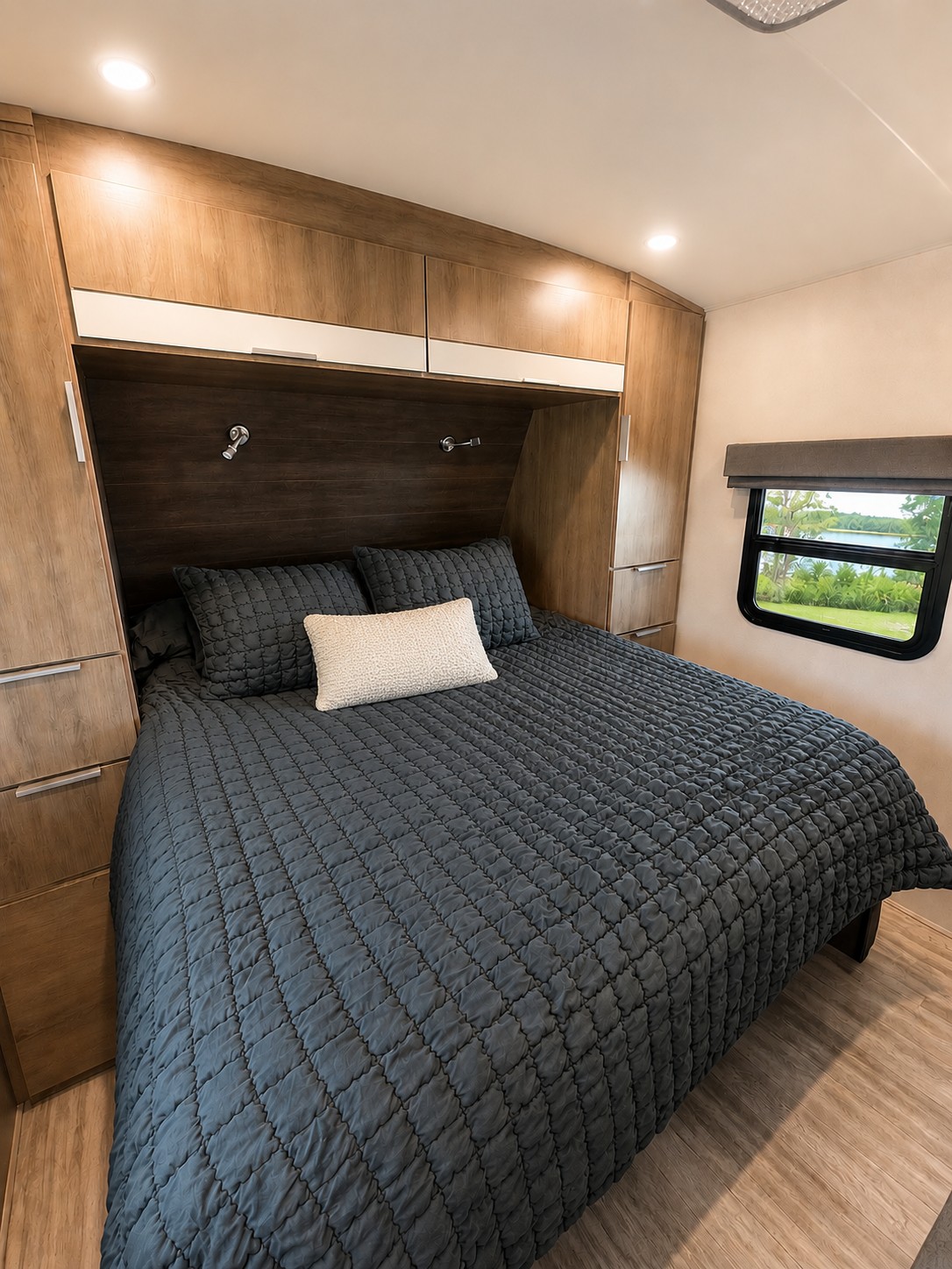 This RV rental's cozy bedroom features a dark gray quilted bed, light wood cabinets, and a window with a scenic view.. Grand Design Imagine 2021