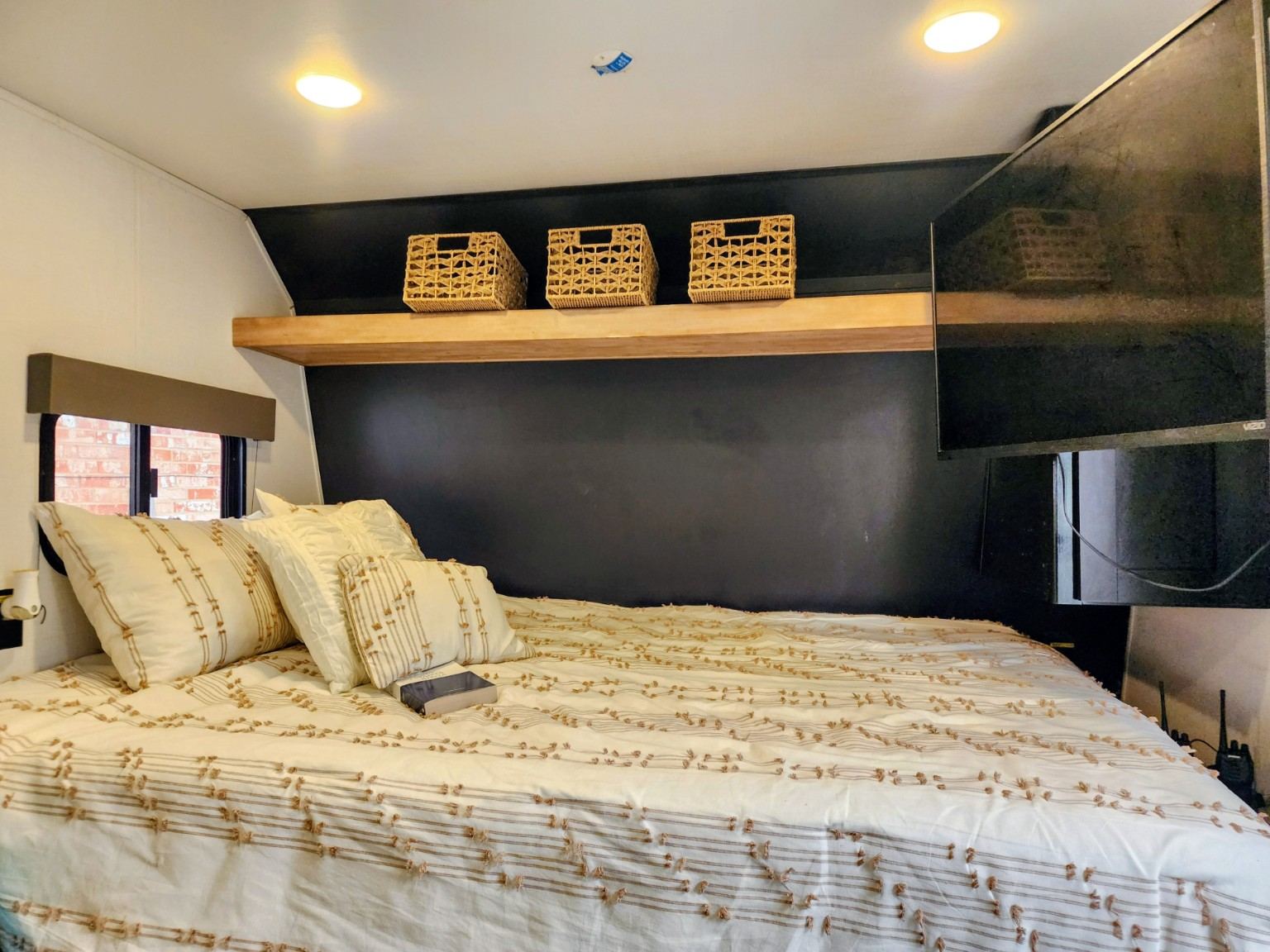 Cozy RV rental bedroom: white/beige bed, dark accent wall, wood shelf with woven baskets, and a wall-mounted TV.. Keystone RV Springdale 2022