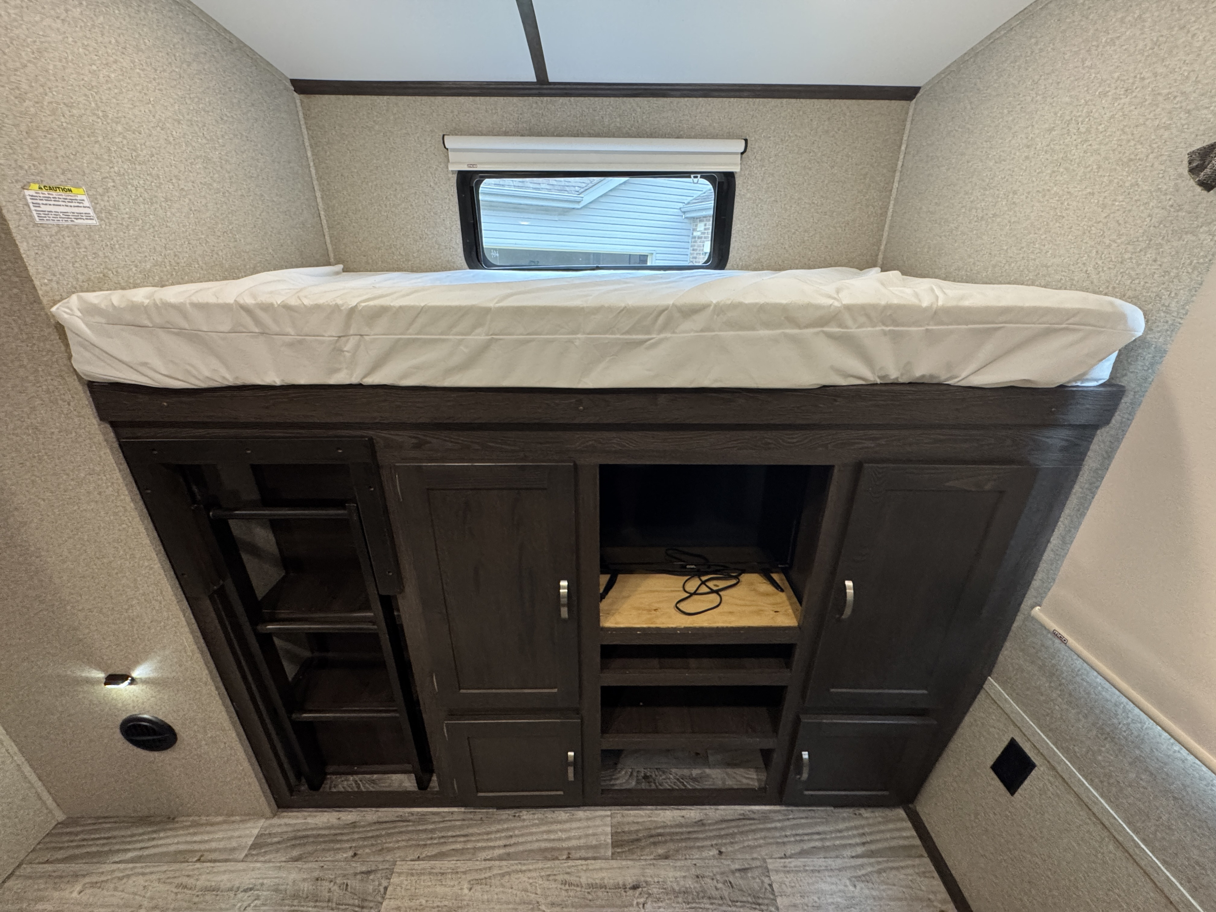 RV rental interior: a comfy bunk bed atop dark built-in cabinets, with a TV, ladder, window, and caution sign visible.. Keystone Outback 2019