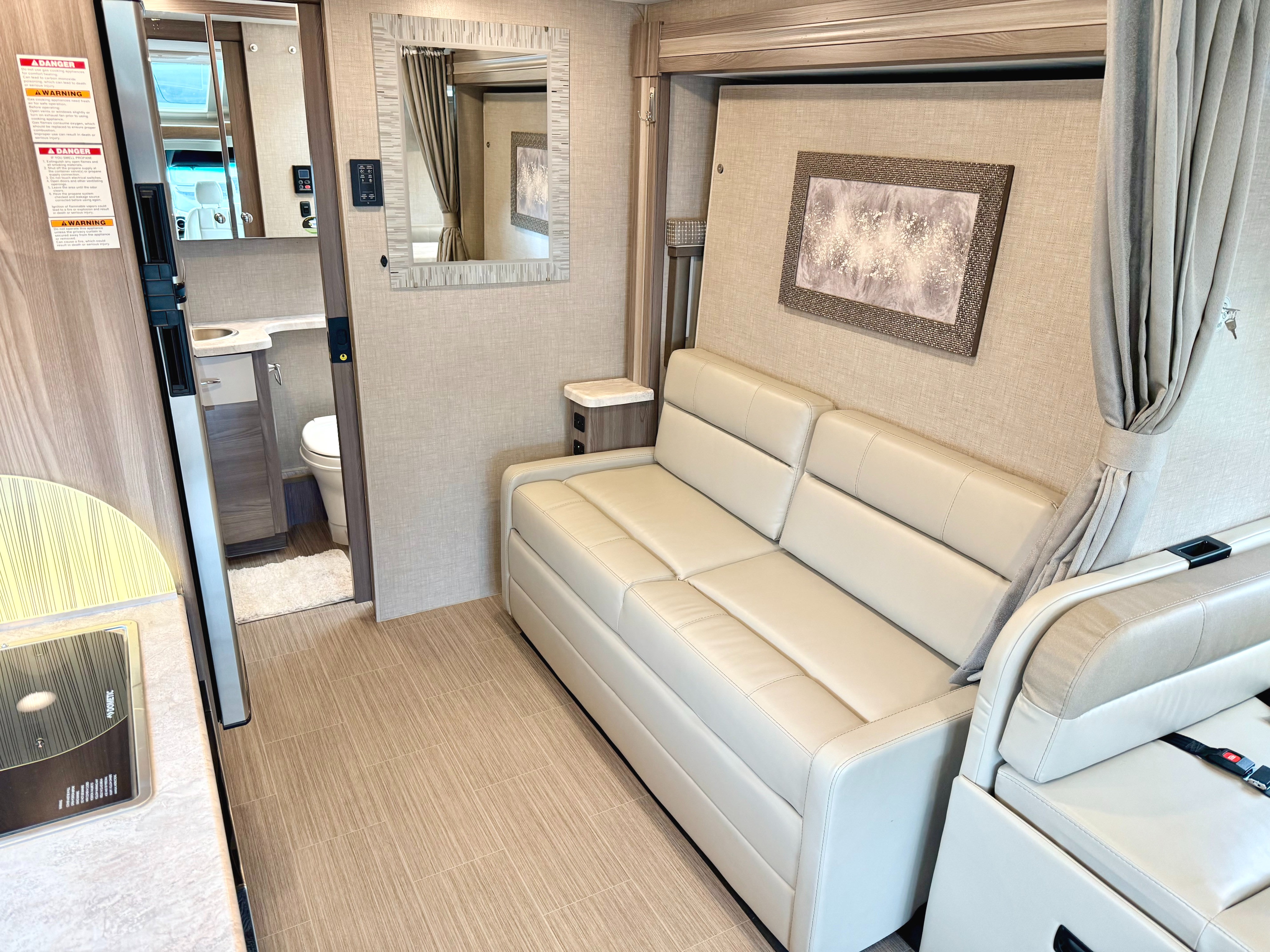 Modern RV rental interior with a light sofa, wall art, mirror, and glimpse of the kitchen and bathroom.. Thor Tiburon 2023