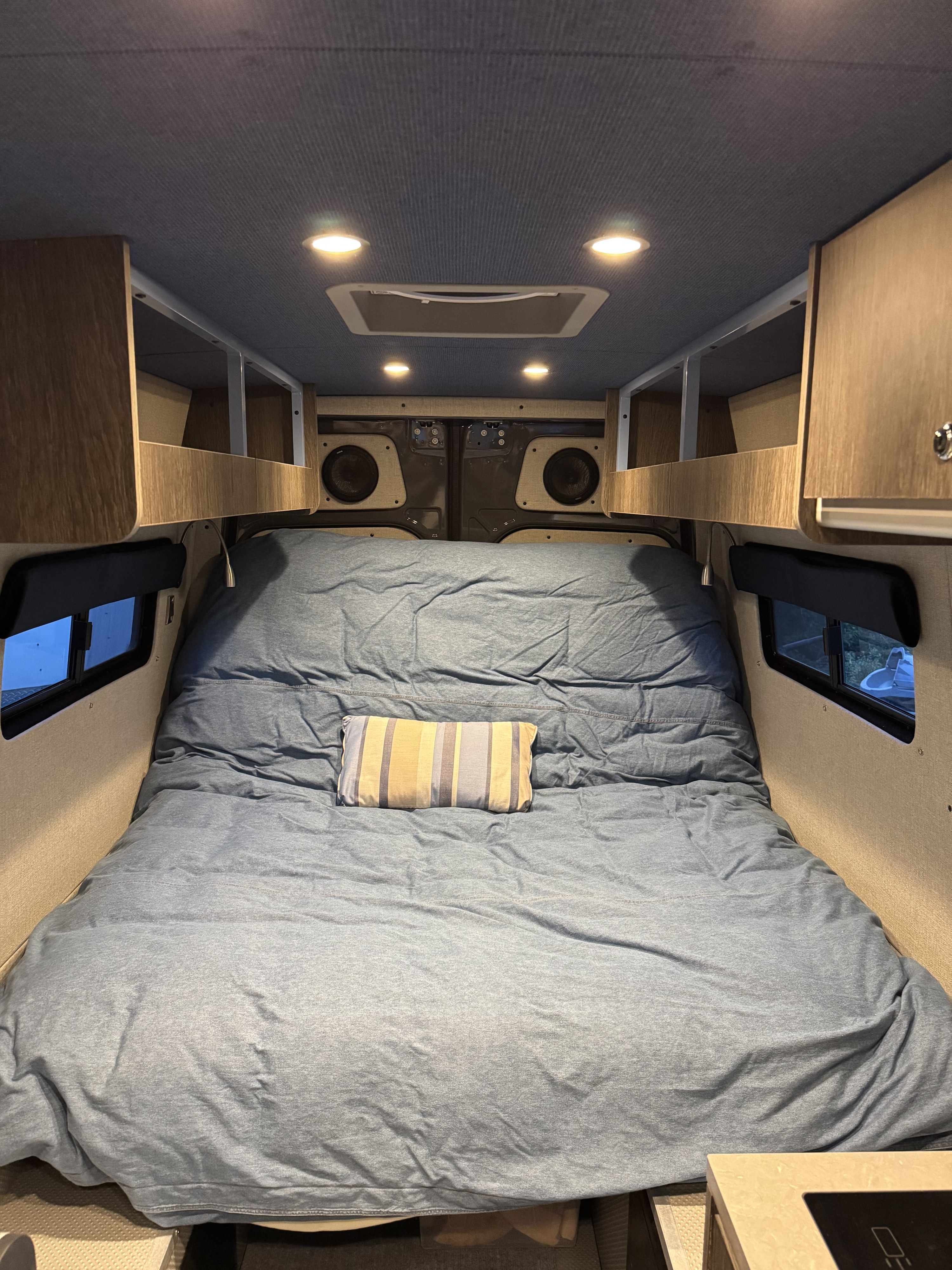 Cozy RV rental camper van interior. A bed with blue bedding, striped pillow, overhead storage, lights, and side windows.. Mercedes Sprinter 2020