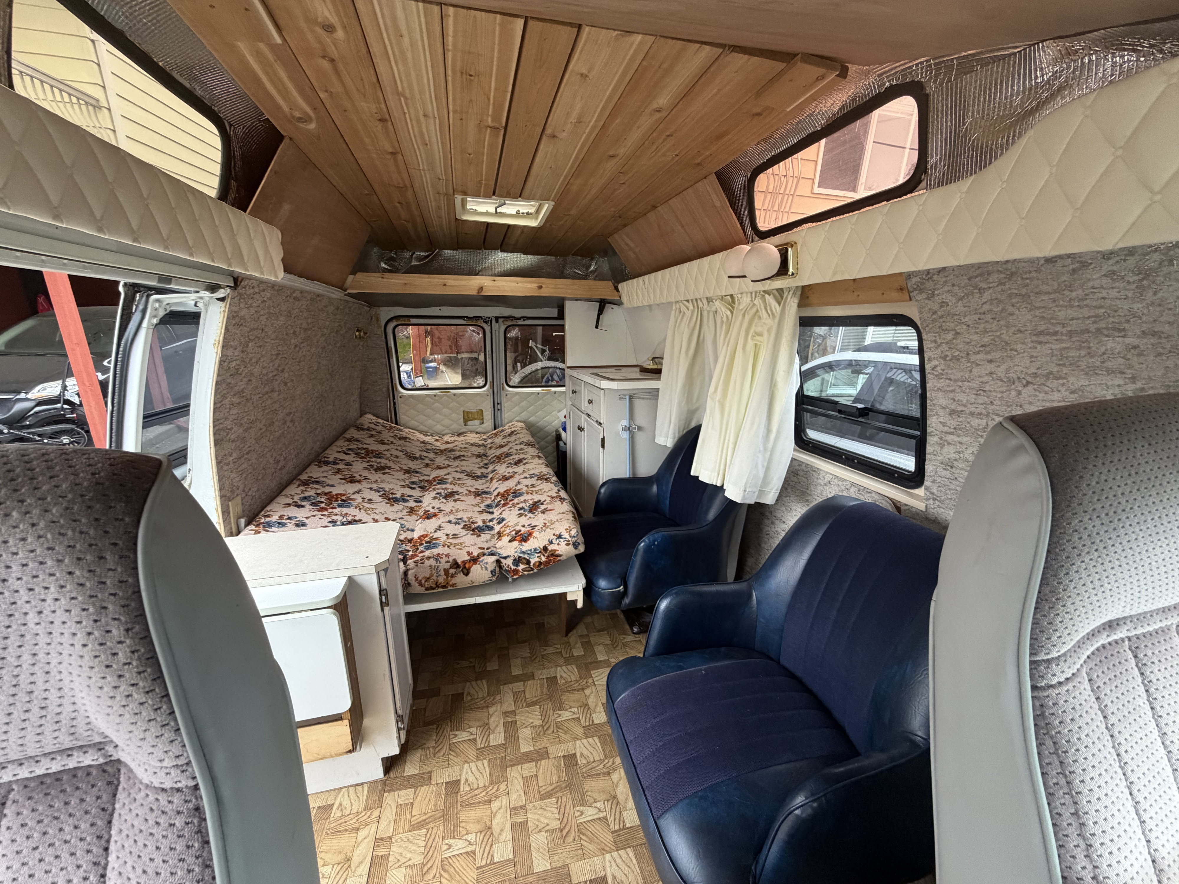 View of a cozy RV rental interior with a wooden ceiling, comfy bed, blue seats, and a compact kitchenette area.. Chevrolet G20 1987