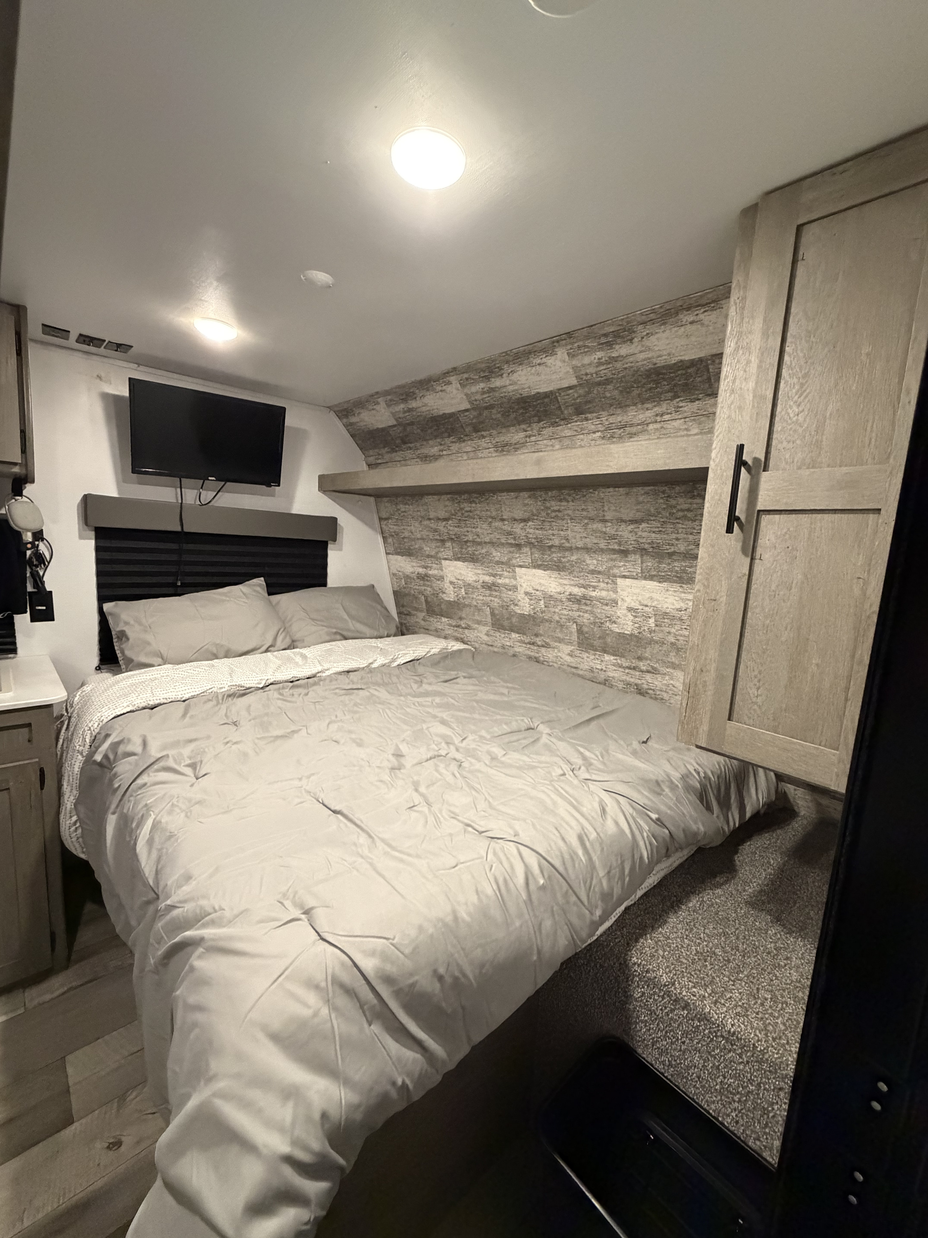 Cozy RV rental bedroom: bed with gray bedding, wall-mounted TV, distressed wood-look walls, and bright ceiling lights.. Keystone RV Springdale 2021