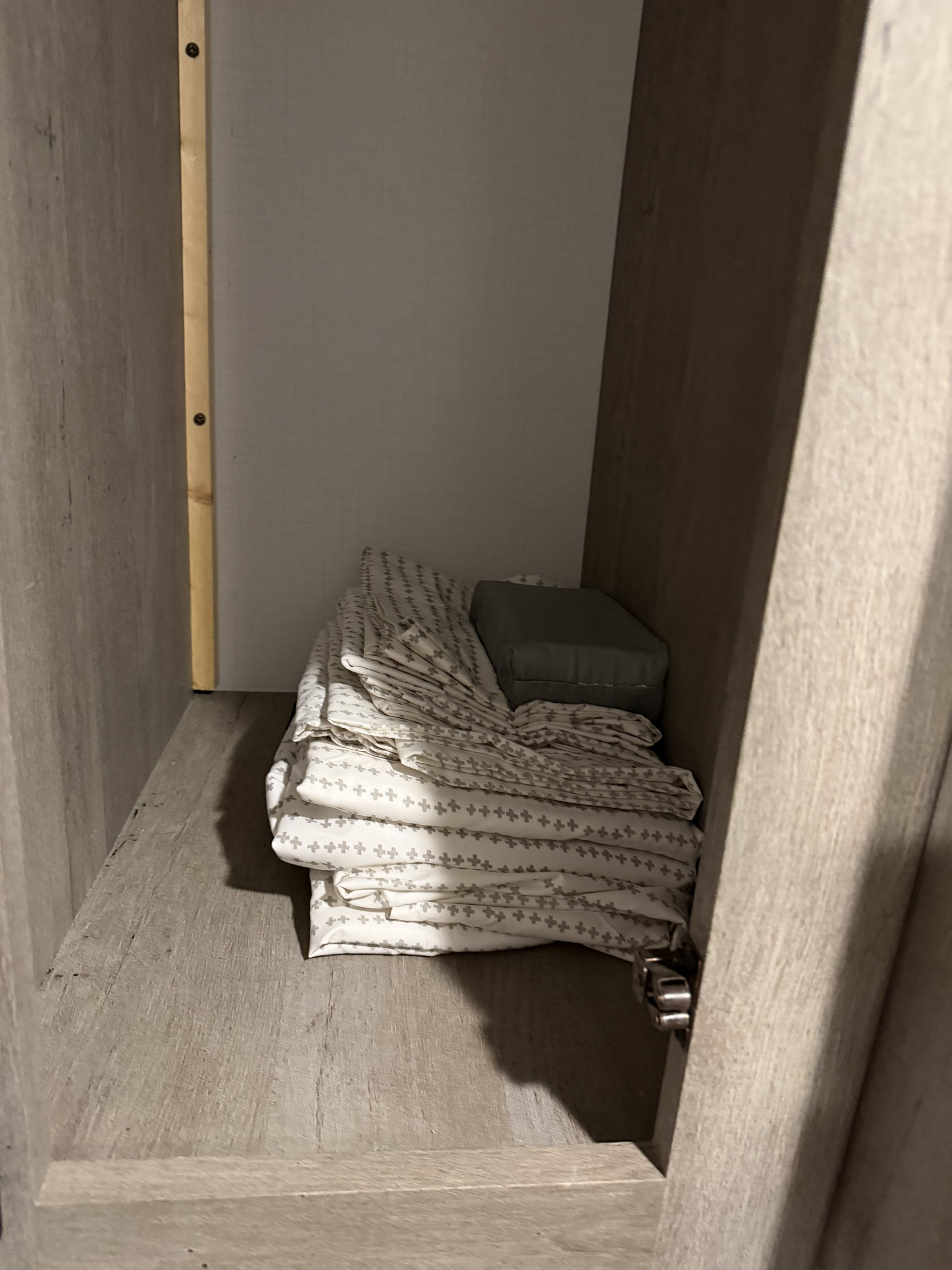 RV rental: Dim wooden cabinet shelf storing neatly folded white sheets with gray cross patterns and a gray cushion.. Keystone RV Springdale 2021
