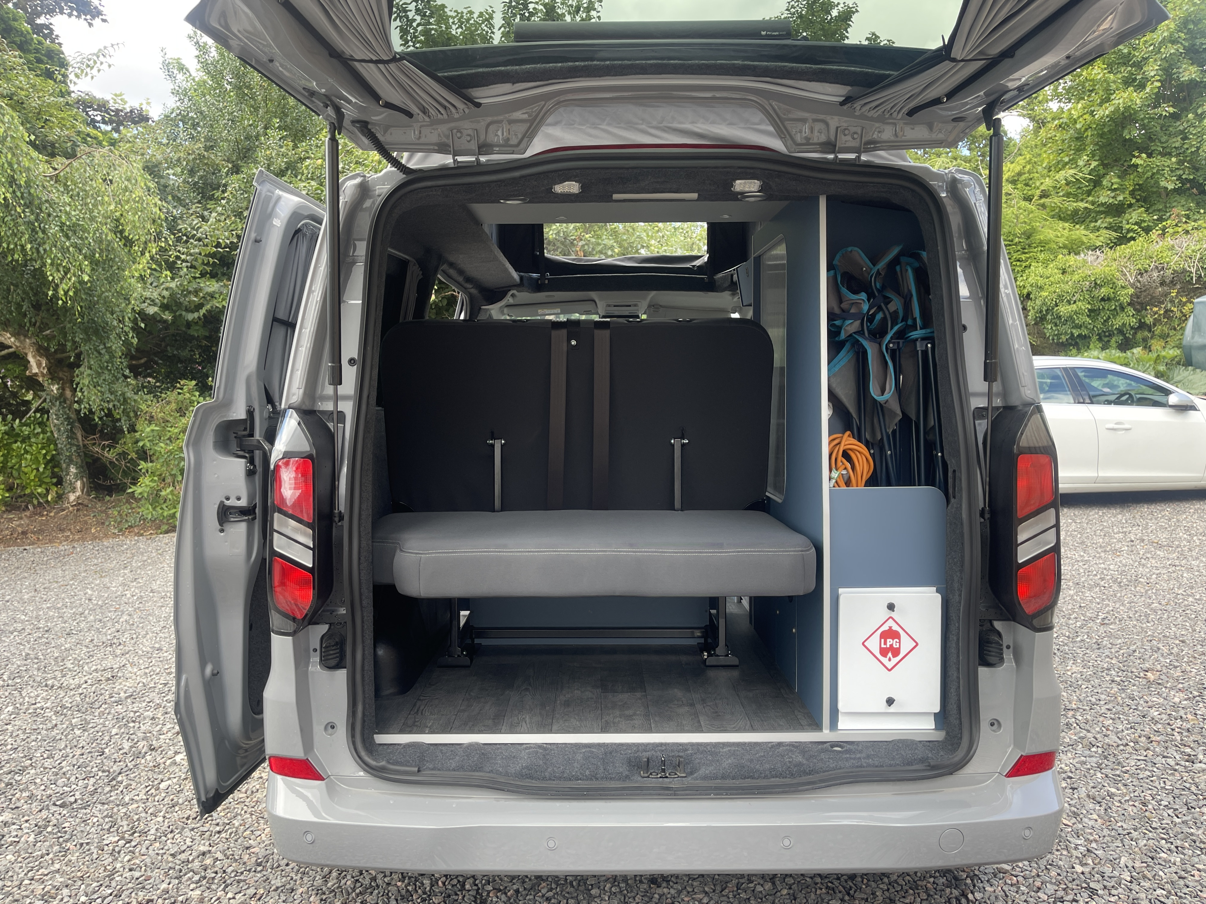 Rear view of an RV rental with its tailgate open, showing a folding bench, storage with camping chairs, and an LPG warning sign inside.. Ford Transit Custom 2+2 berth Pop Top, Automatic 2026